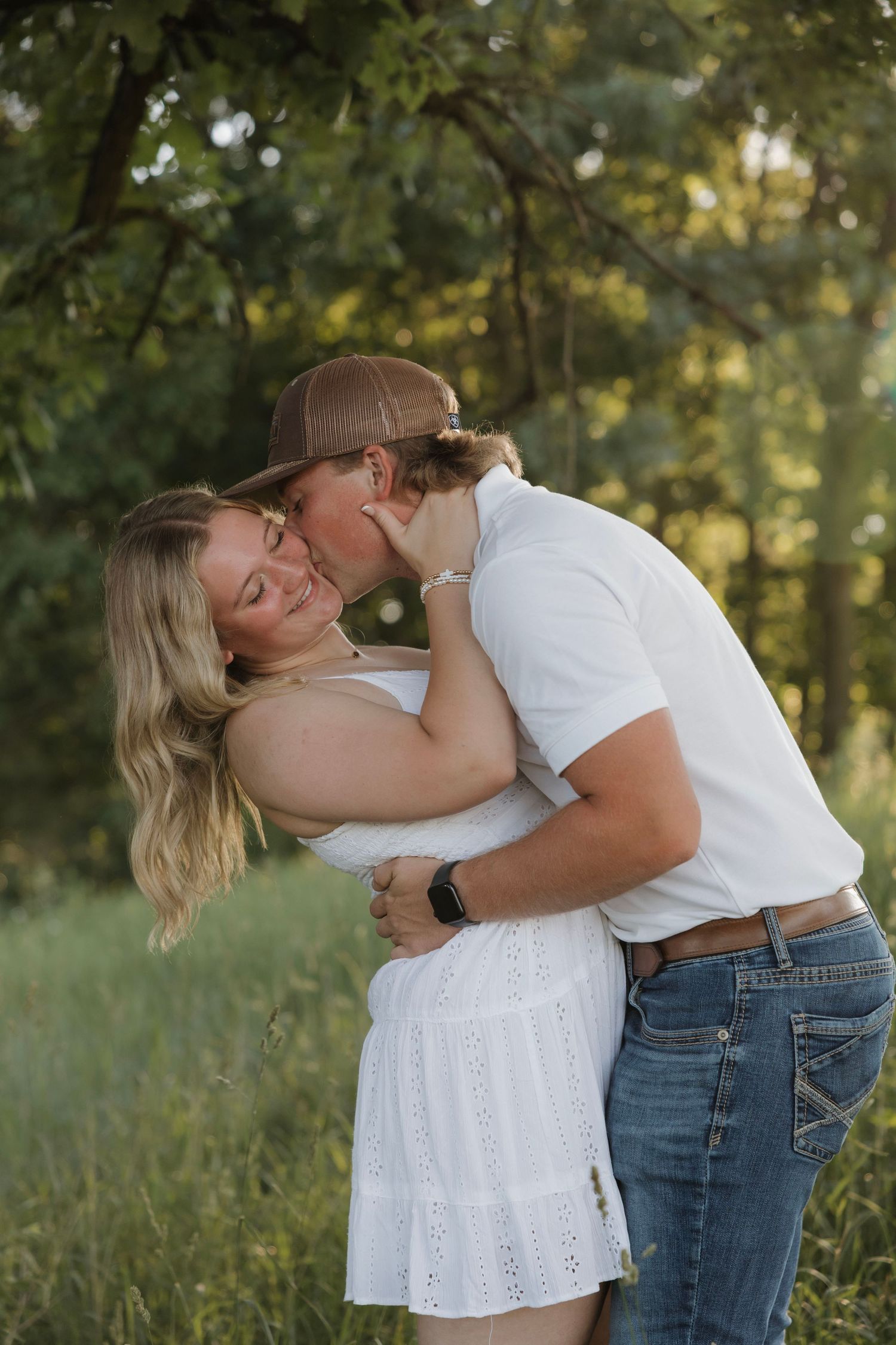 Couple embracing and kissing in lush green park, woman in white dress and man in white shirt and jeans.