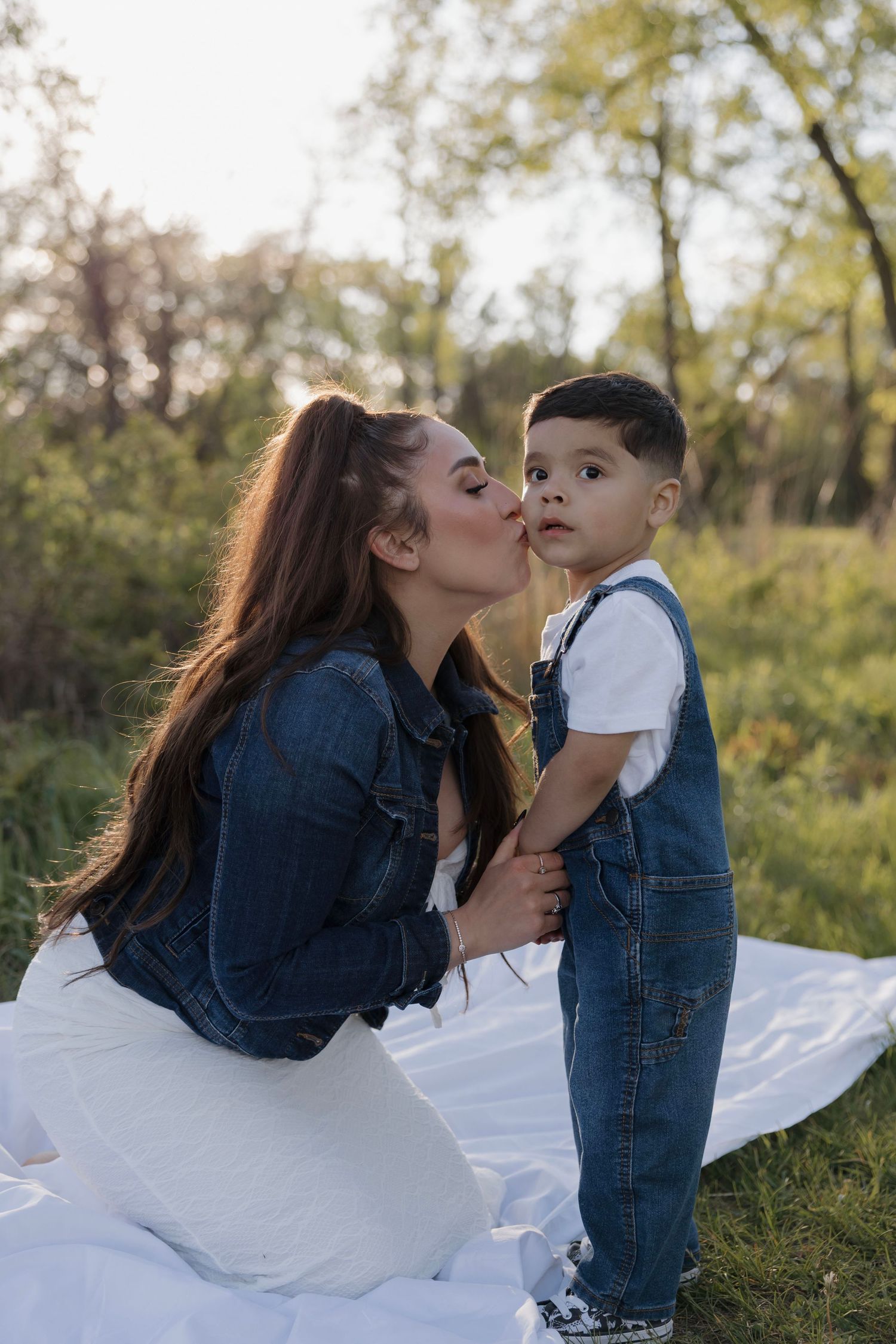 Young mother in denim jacket kissing her toddler son on the cheek outdoors on a blanket.