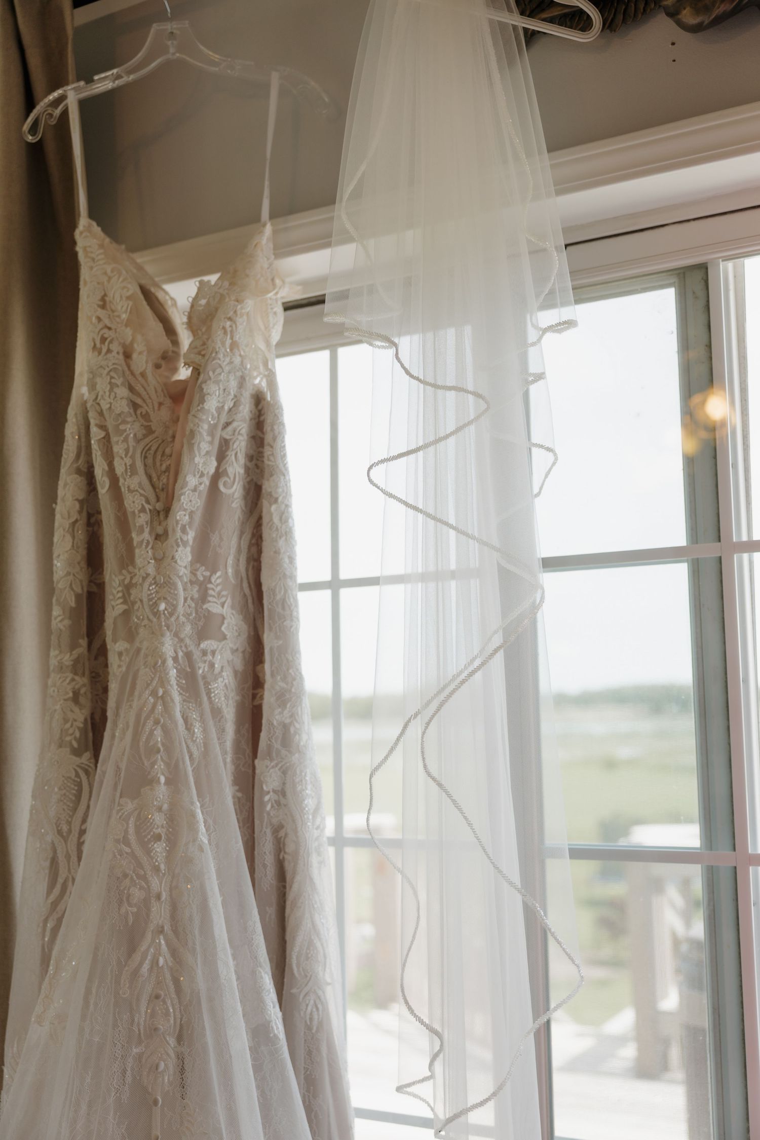 Elegant lace wedding dress and flowing veil hanging by a bright window with scenic outdoor view.