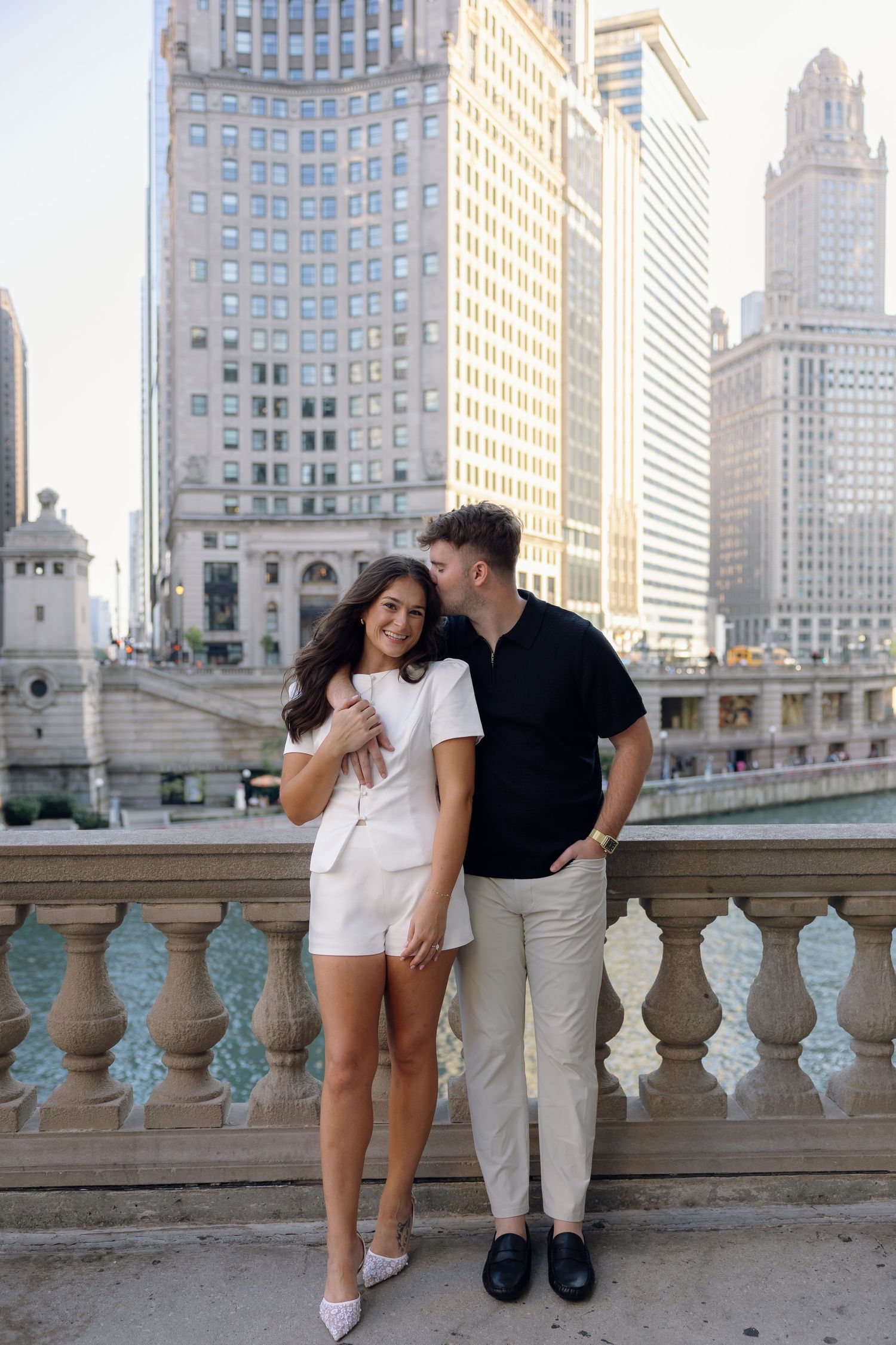 A couple poses romantically on a bridge with Chicago skyline buildings in the background on a sunny day.