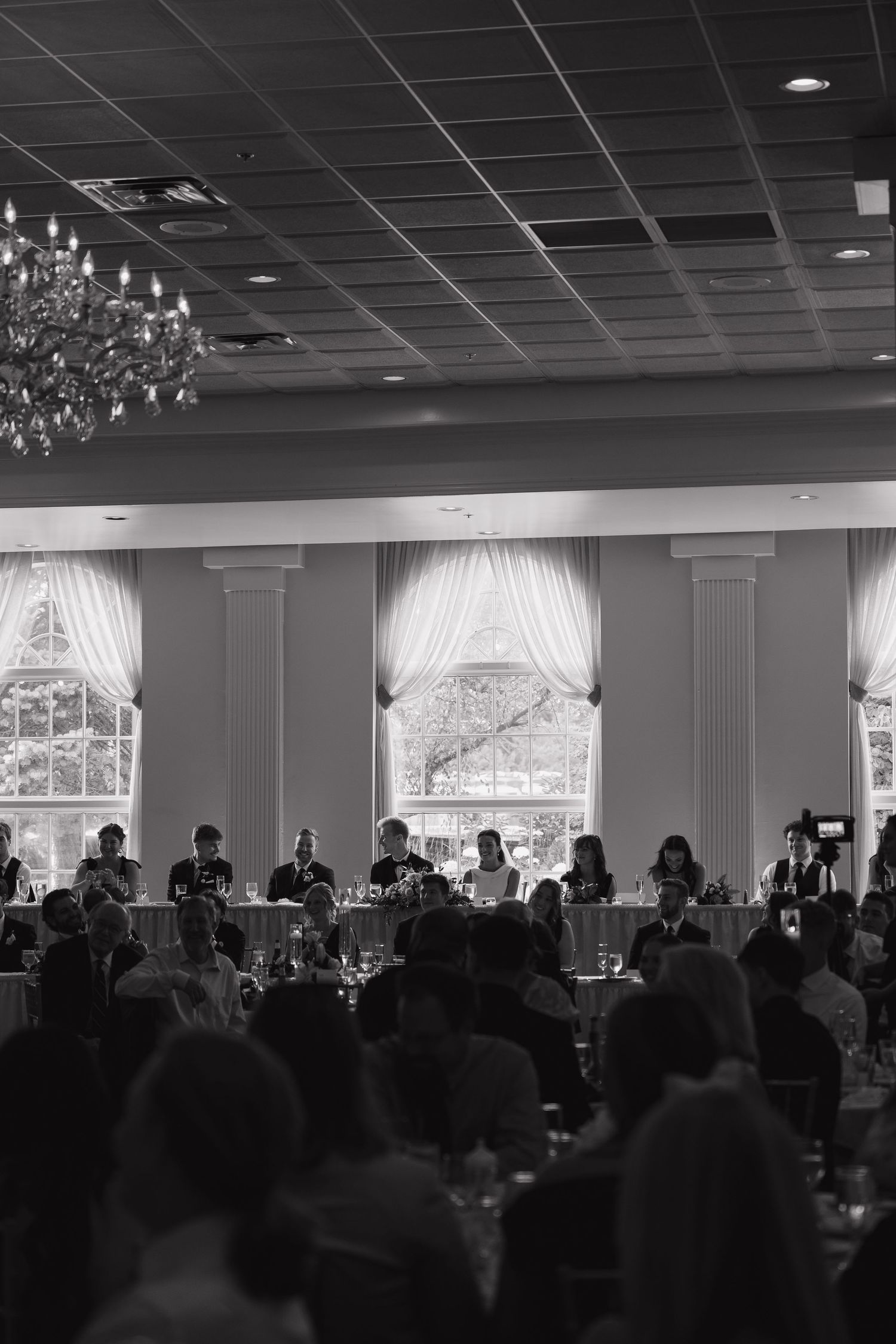 Black and white photo of elegant wedding reception banquet hall with chandelier, draped windows, and guests seated at tables.