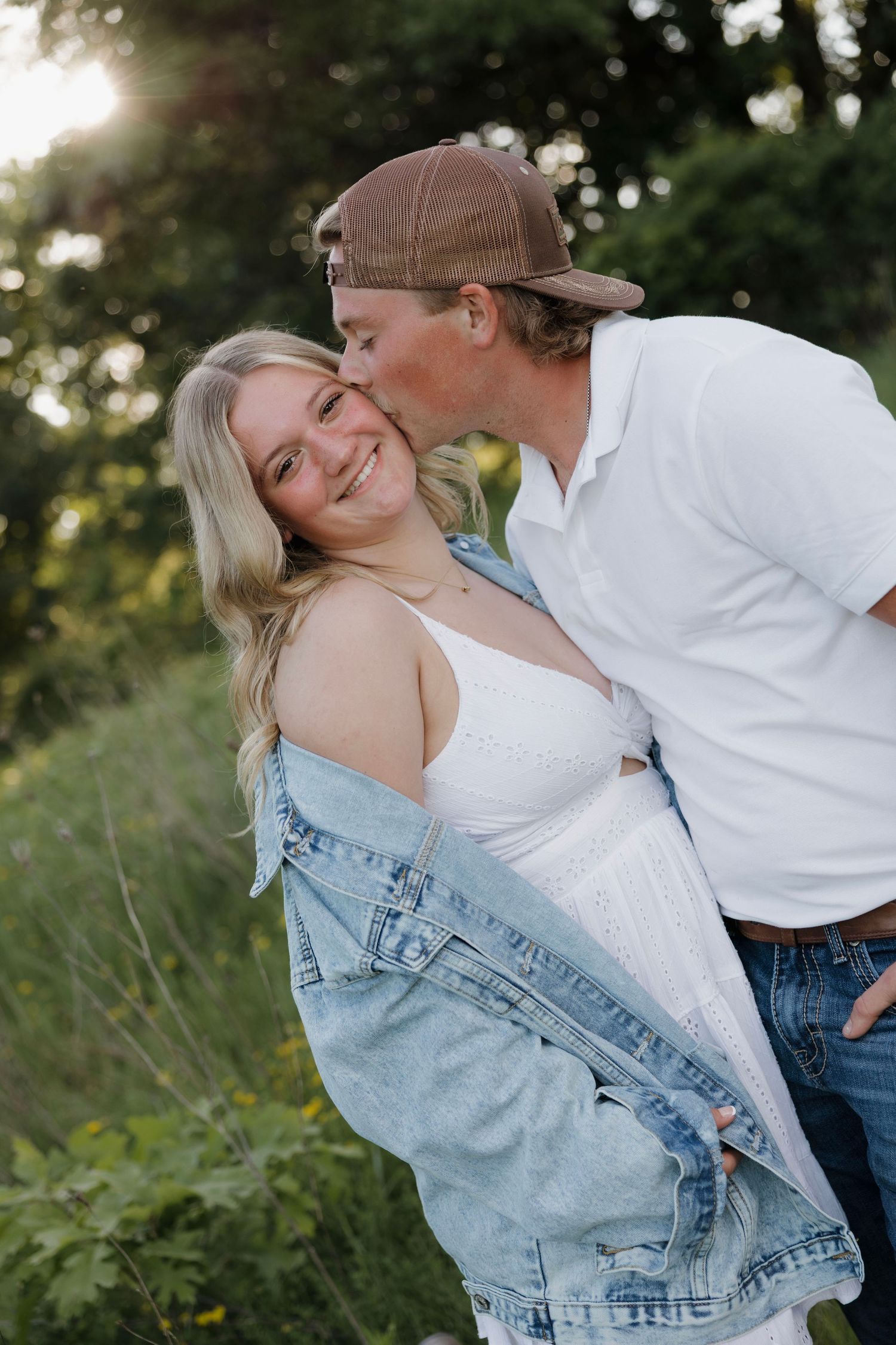 Man kissing smiling blonde woman on cheek outdoors in sunlit green field, woman wearing denim jacket.