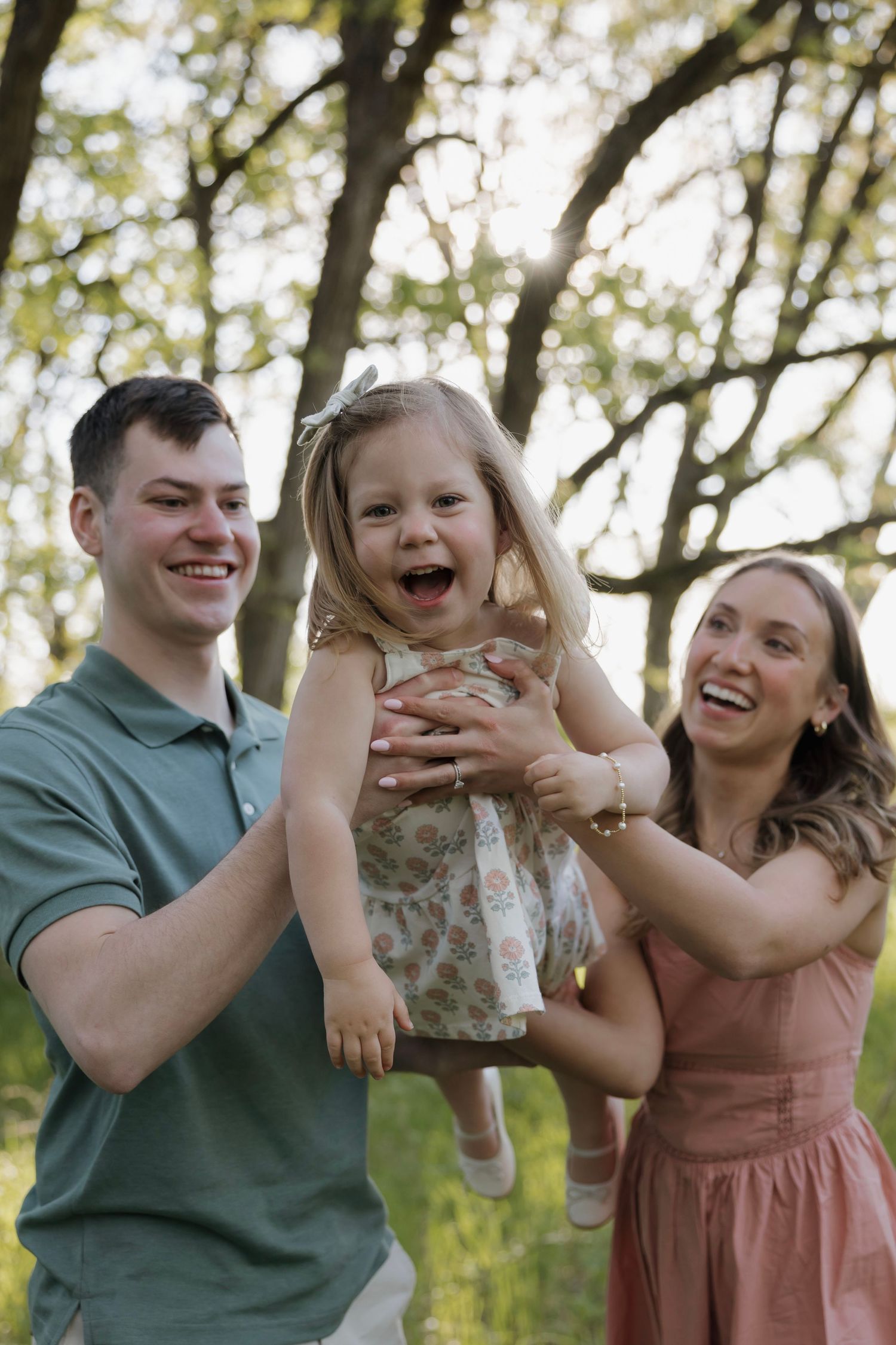 Happy young family with parents lifting laughing toddler girl outdoors in sunny park setting.