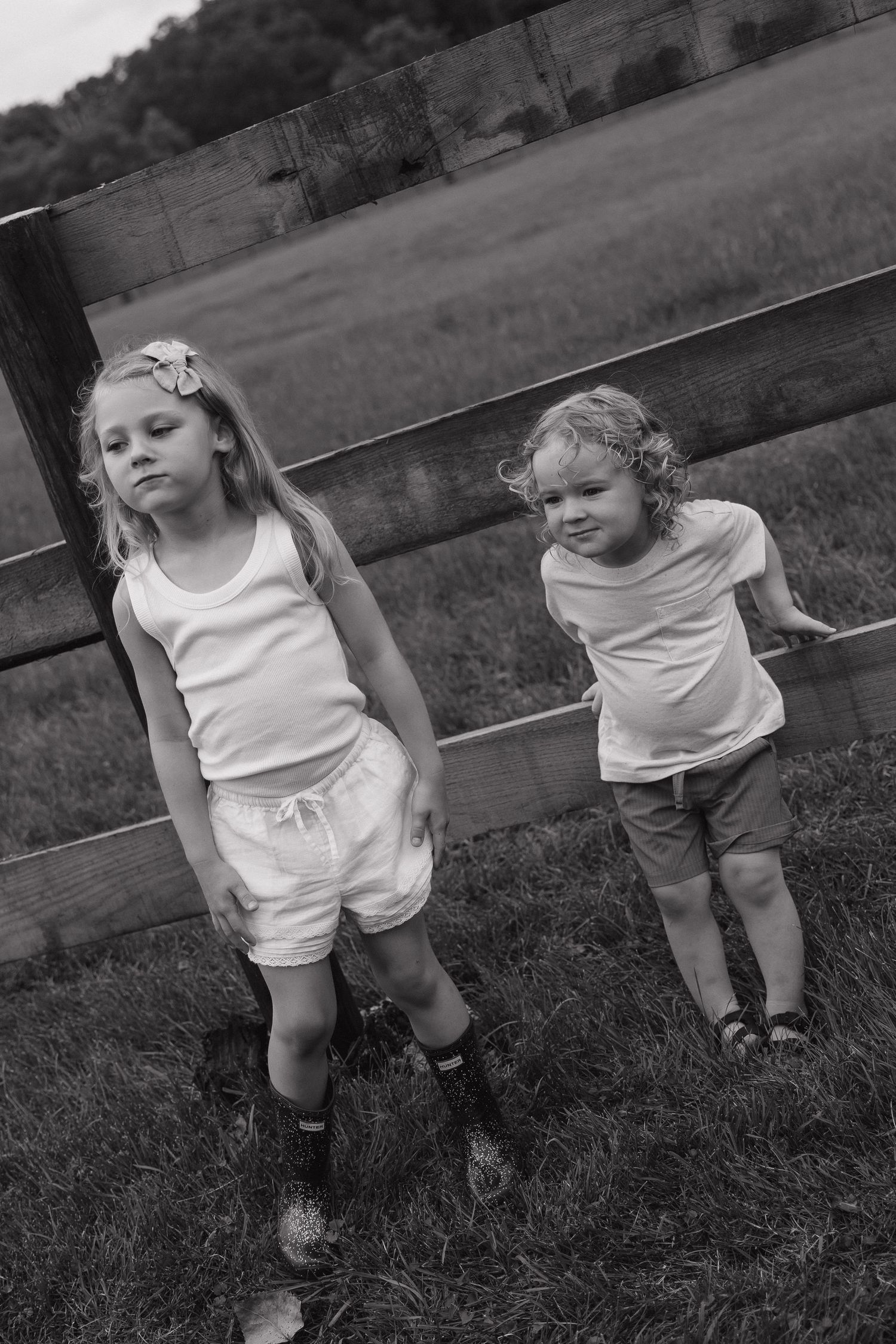 Two young children standing by wooden fence in field in black and white photo.