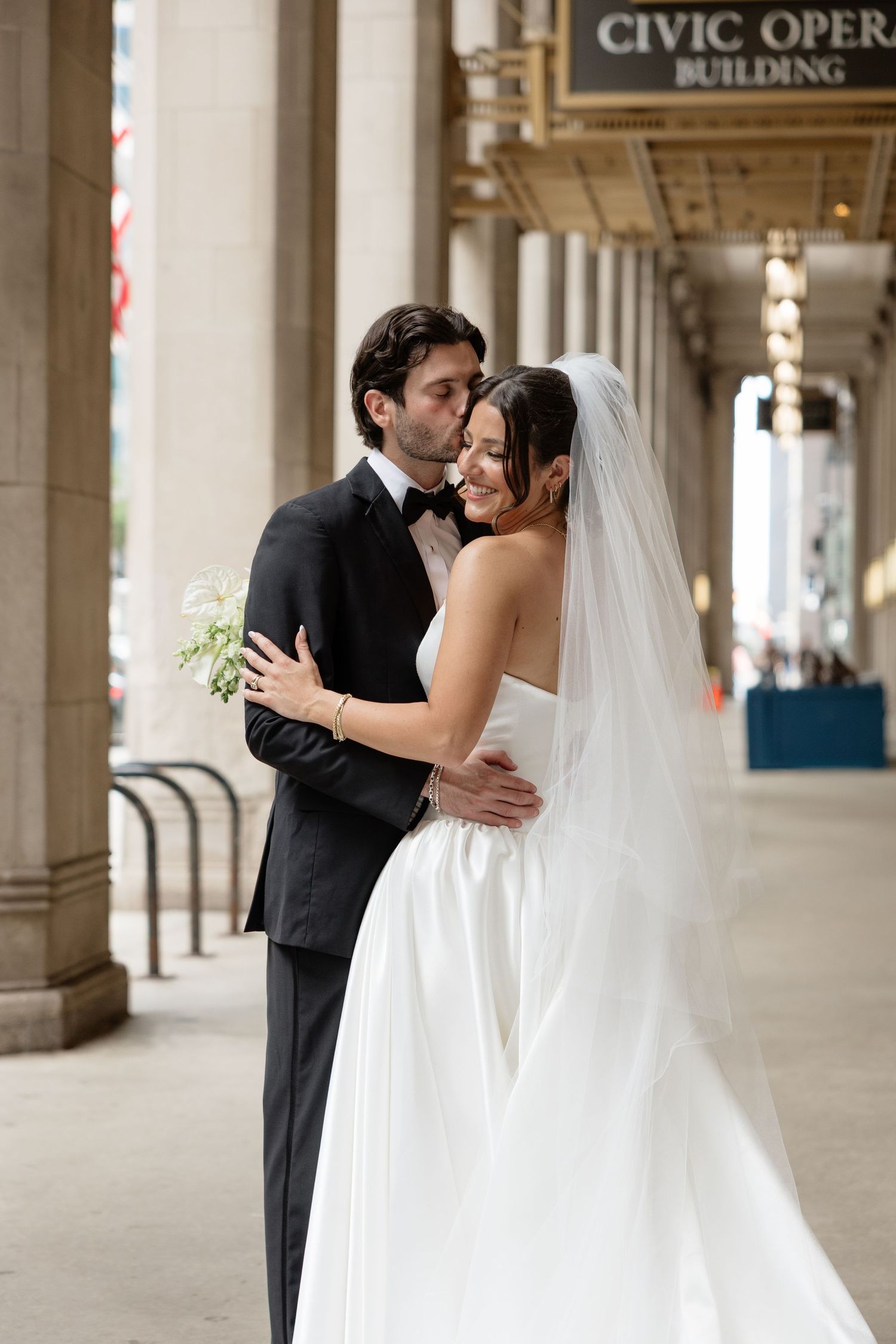 Bride and groom embrace outside Chicago Civic Opera Building, she in white gown with veil, he in black tuxedo.