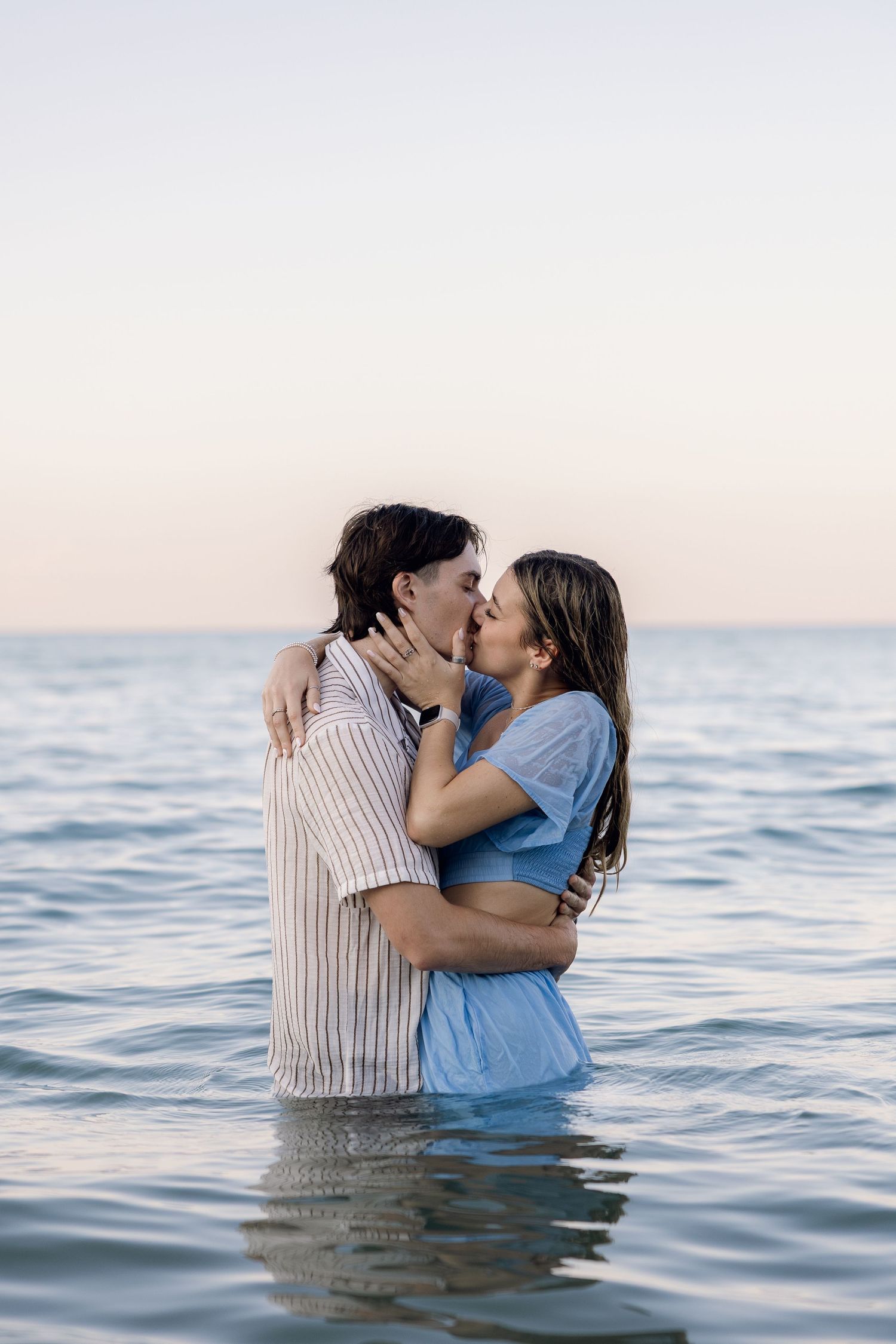 Romantic couple kissing while standing in ocean water at sunset, wearing casual summer clothes.
