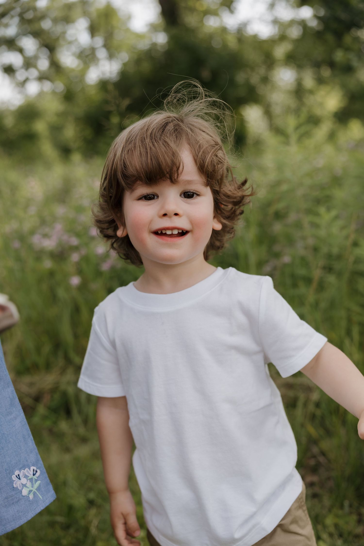 Happy curly-haired toddler boy in white t-shirt smiling in a green meadow.