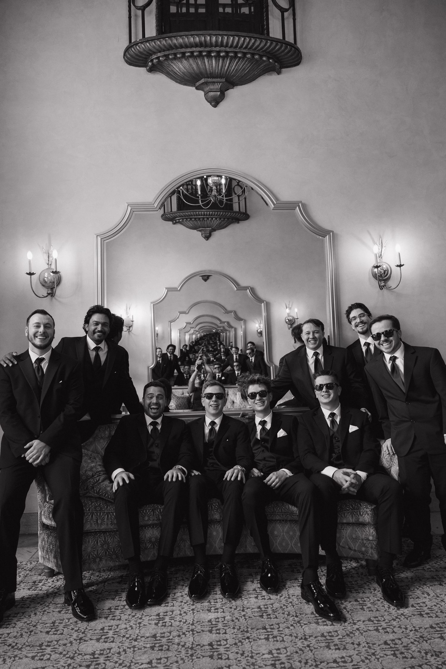 Black and white photo of groomsmen in suits and sunglasses posing on a sofa in an elegant wedding venue.