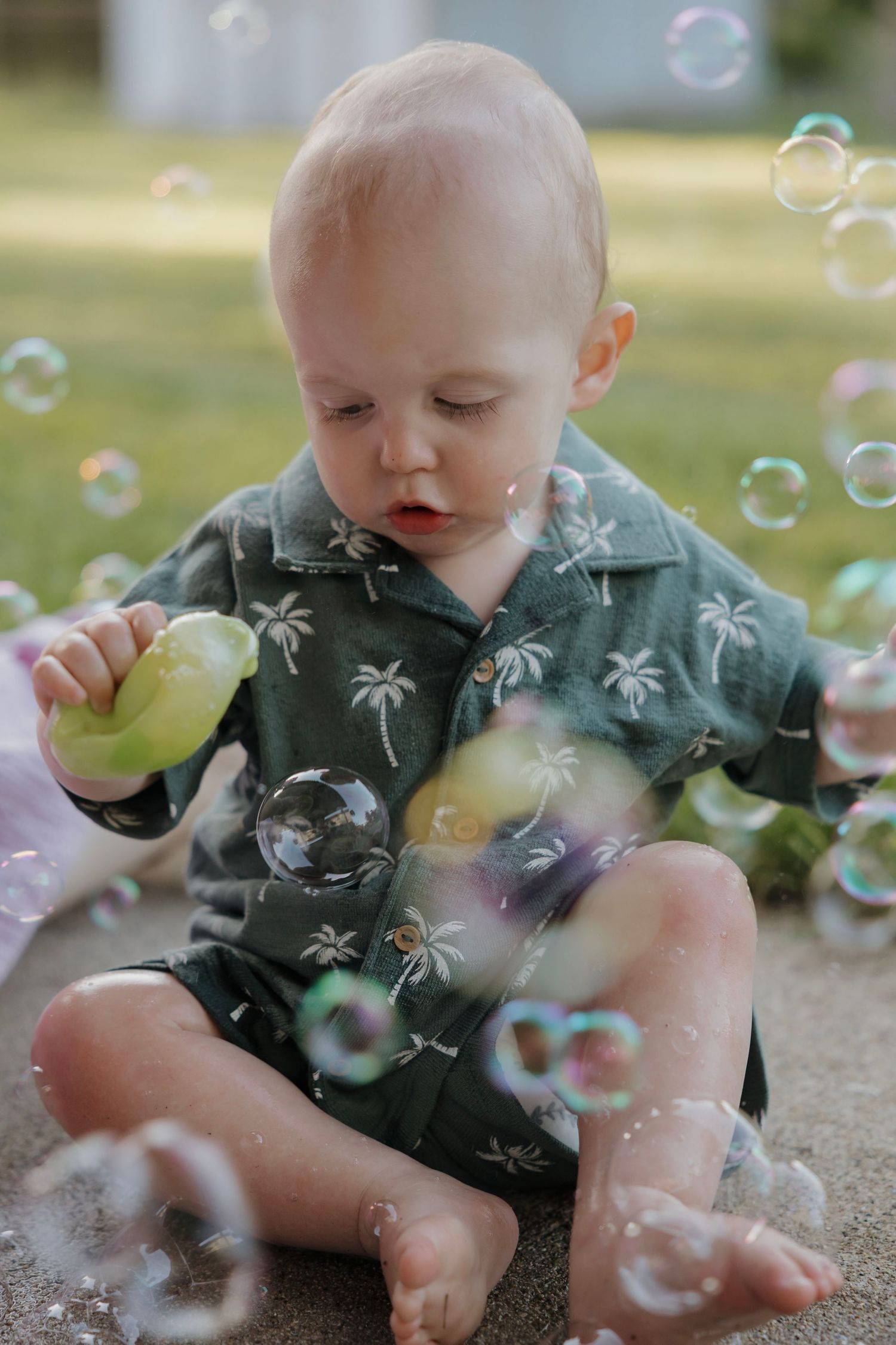 Baby in green palm tree shirt sitting on pavement surrounded by floating bubbles.