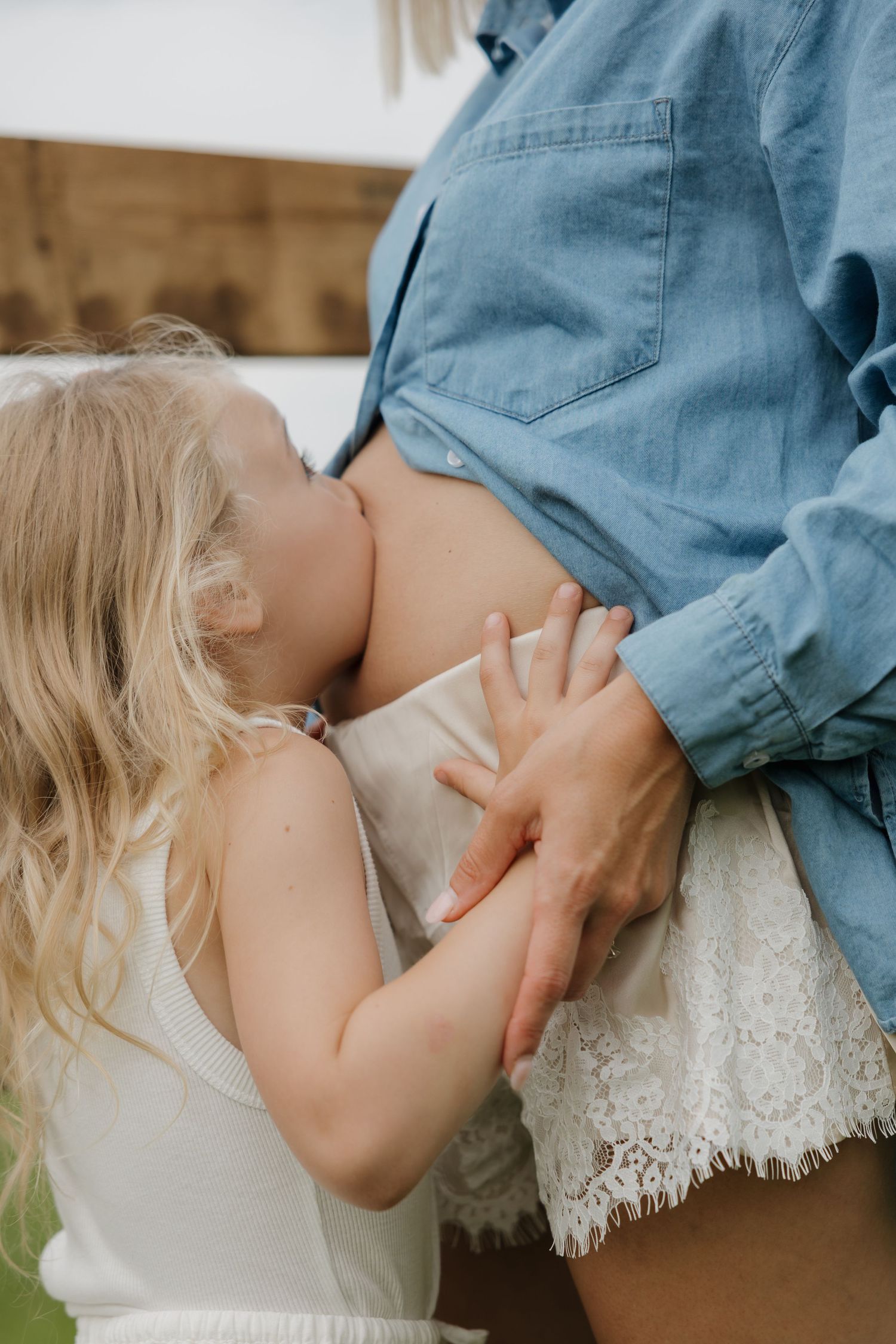 Young blonde girl kissing mothers pregnant belly outdoors in natural light.