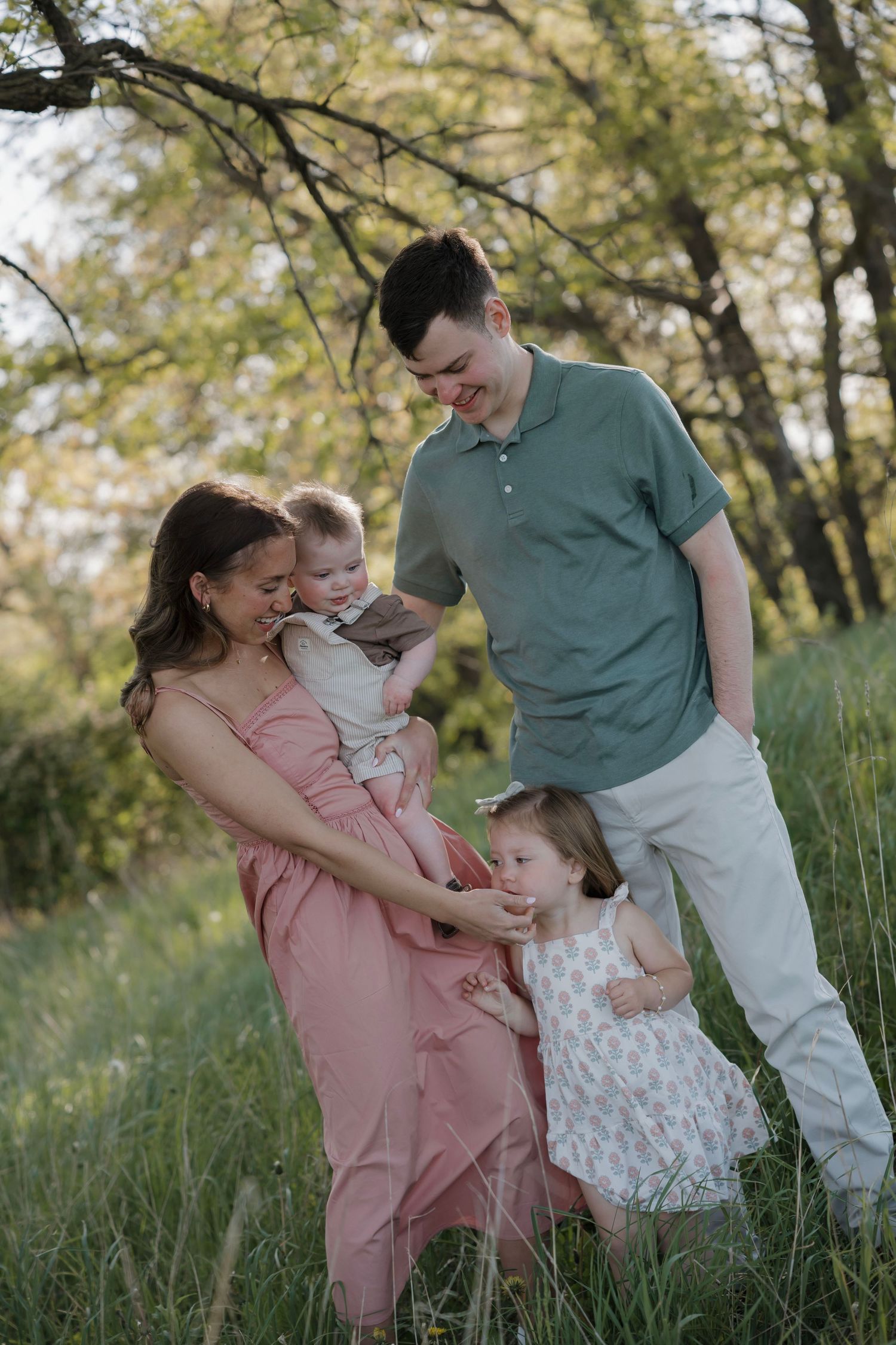 Young family of four with mother in pink dress and father in green polo enjoying time in a meadow.