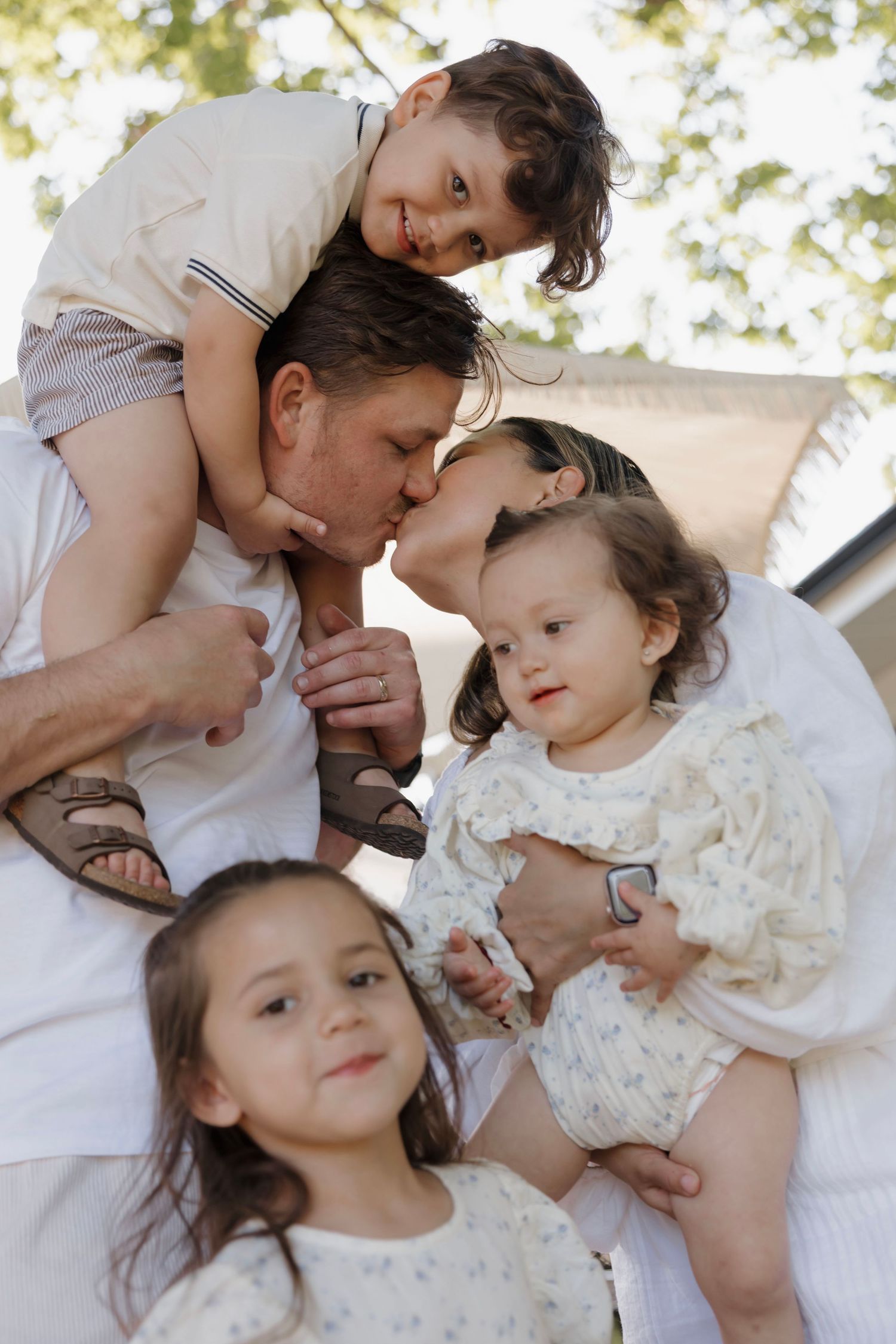Happy family of five with father kissing mother while children surround them outdoors under trees.