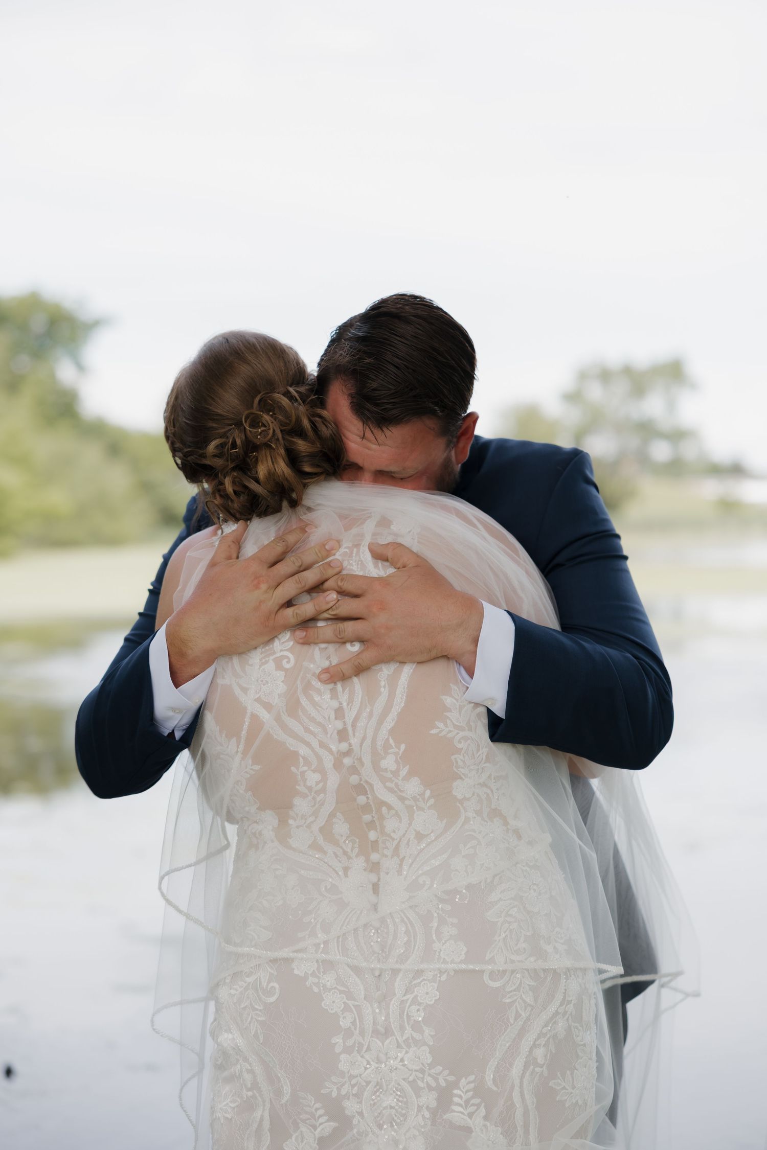 Groom in navy suit embracing bride in lace wedding dress during emotional first look near a serene waterfront.