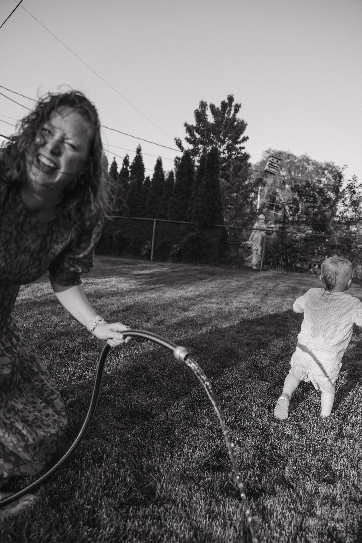 Black and white photo of laughing woman with garden hose as toddler runs away.