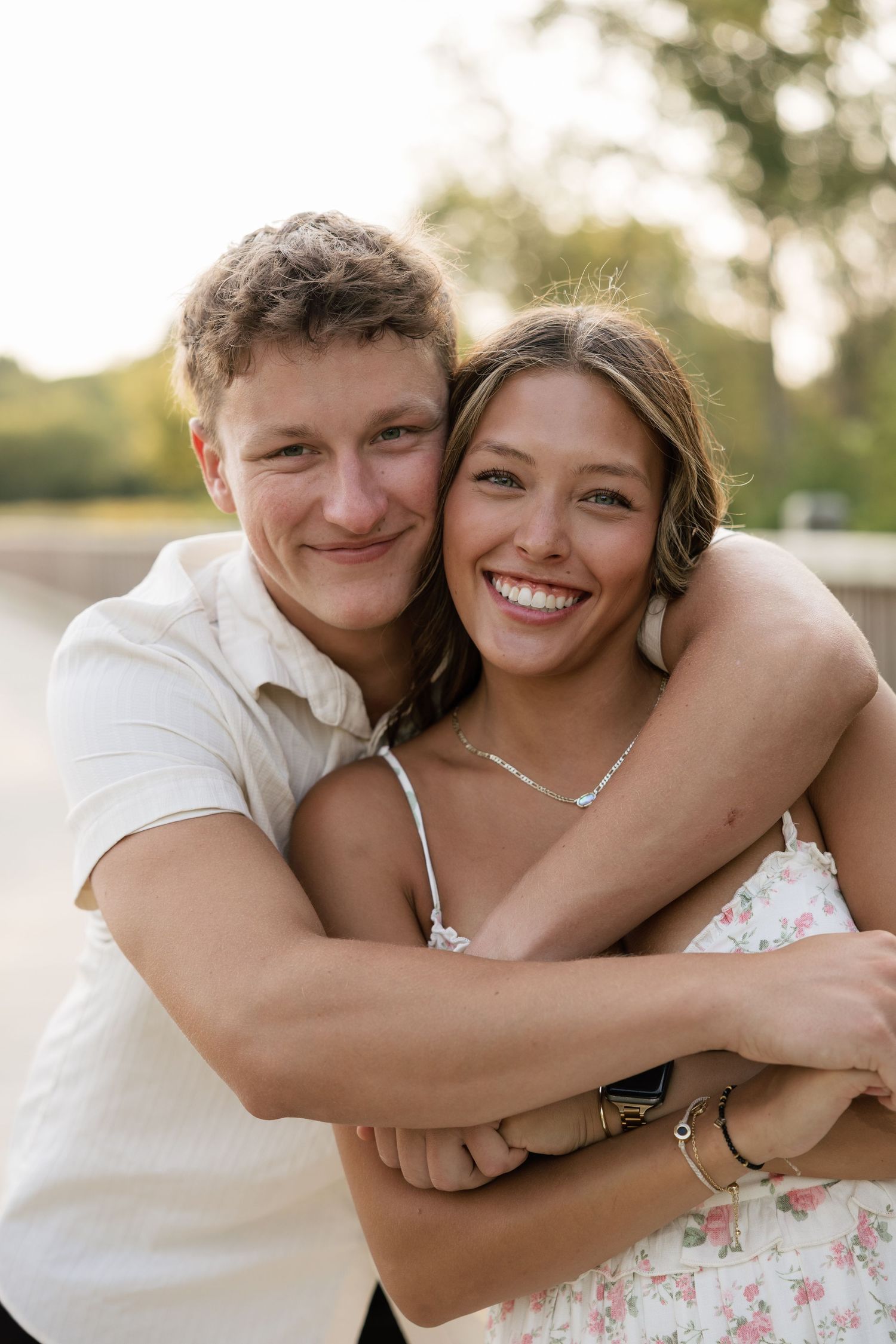 Smiling couple embracing outdoors at golden hour, woman in floral dress laughing joyfully.