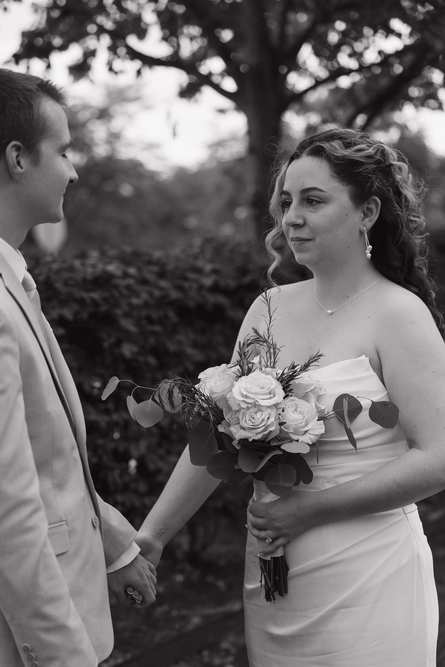 Black and white wedding photo of bride holding rose bouquet and groom holding hands outdoors.