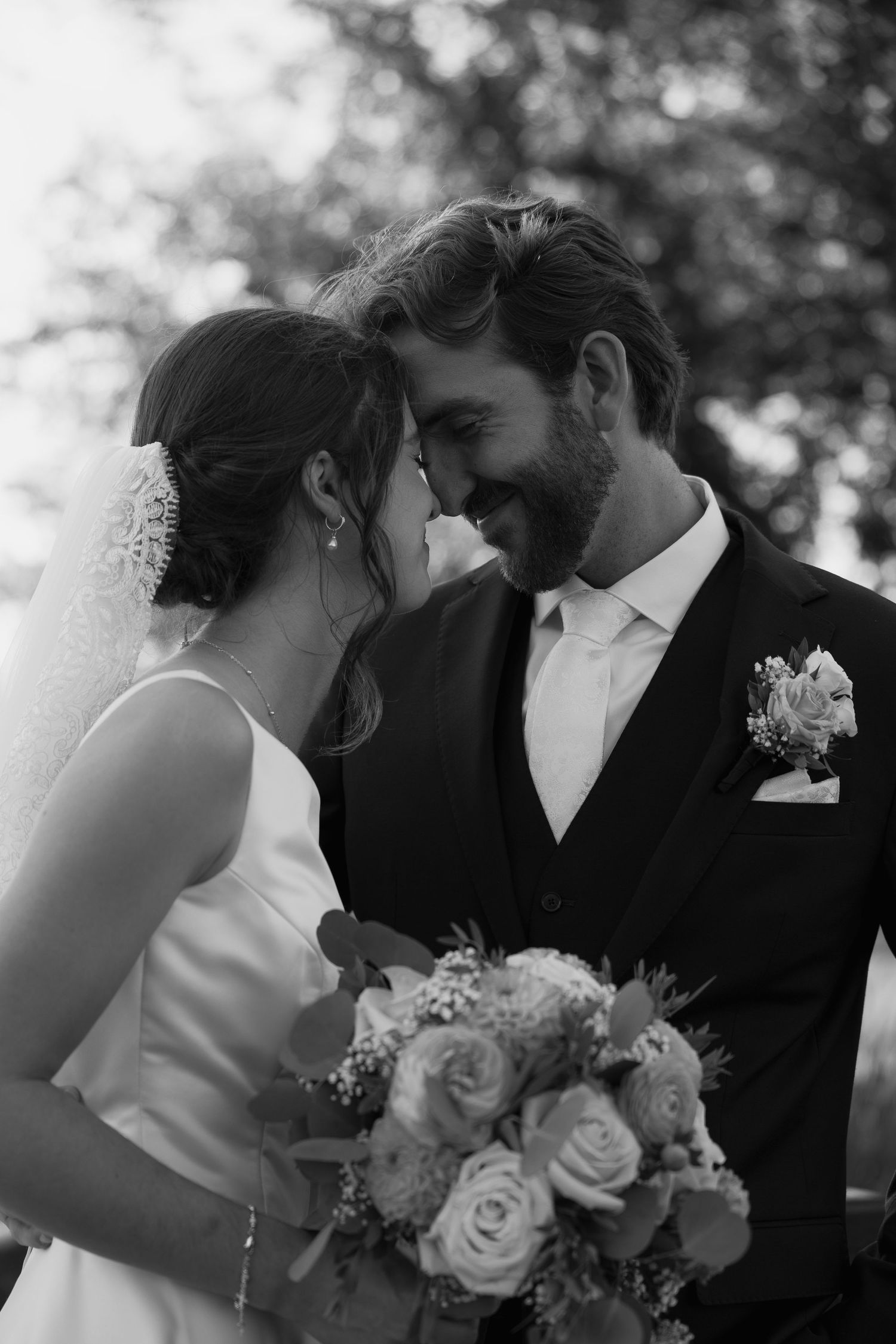 Black and white photo of a smiling bride and groom touching foreheads, bride holding a rose bouquet.