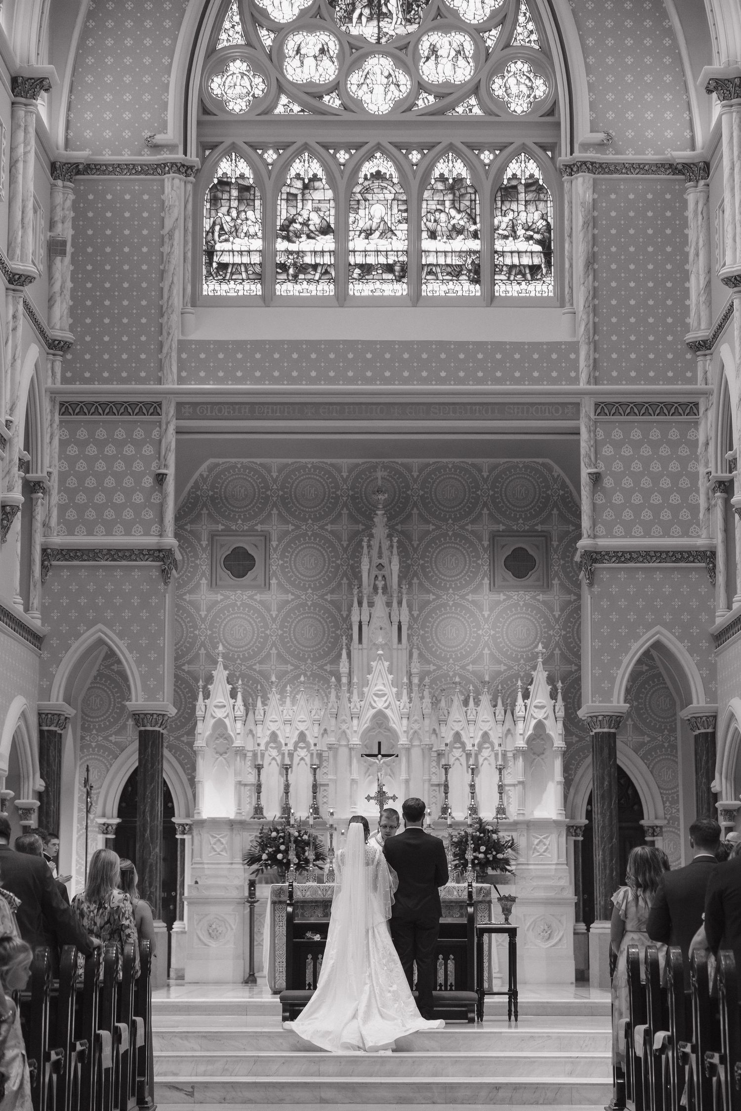 Black and white photo of a bride and groom at the altar of an ornate Gothic church with stunning stained glass windows.
