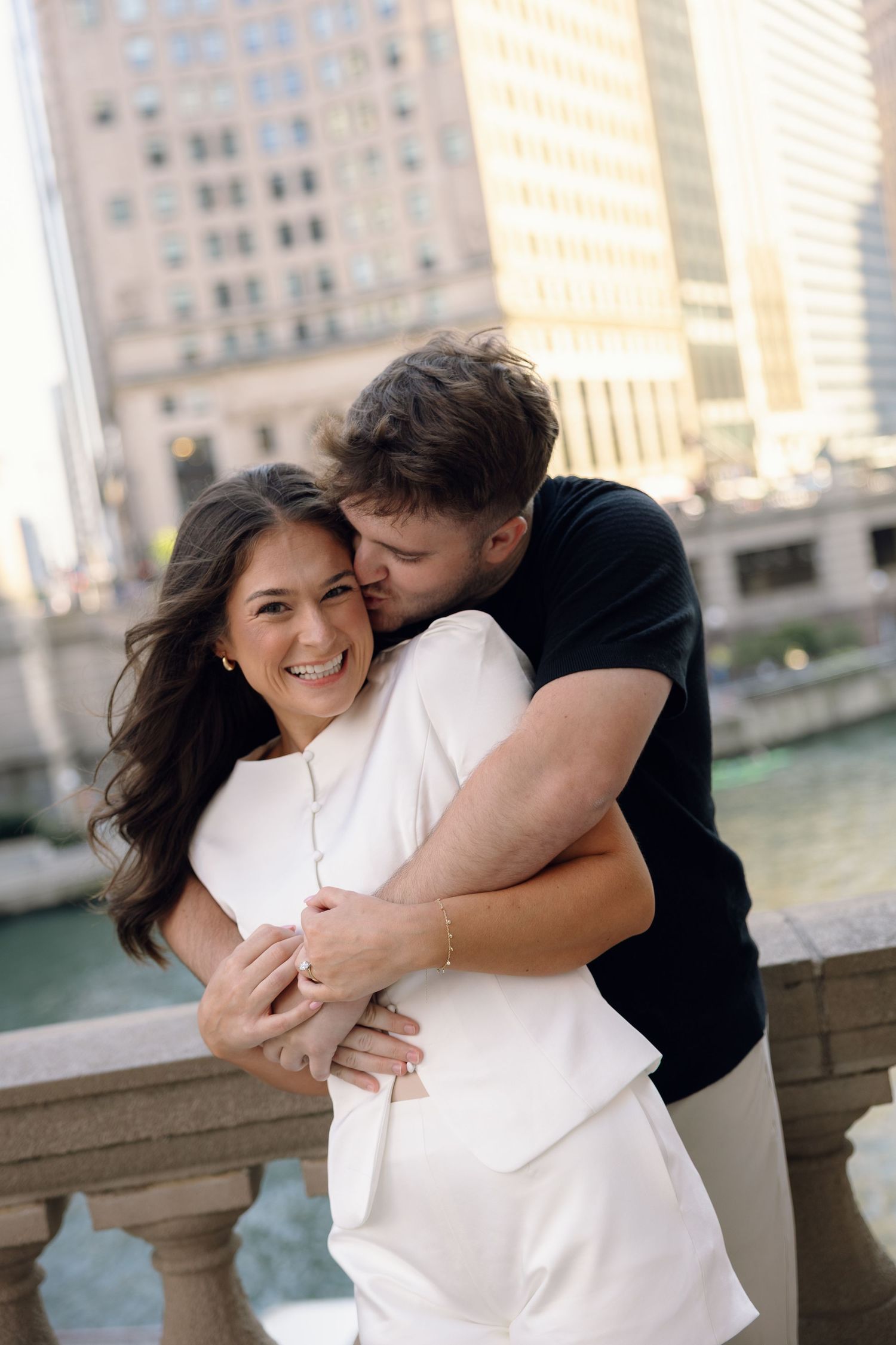 Couple embracing and smiling on a bridge overlooking a city river, woman in white outfit, man in black shirt.