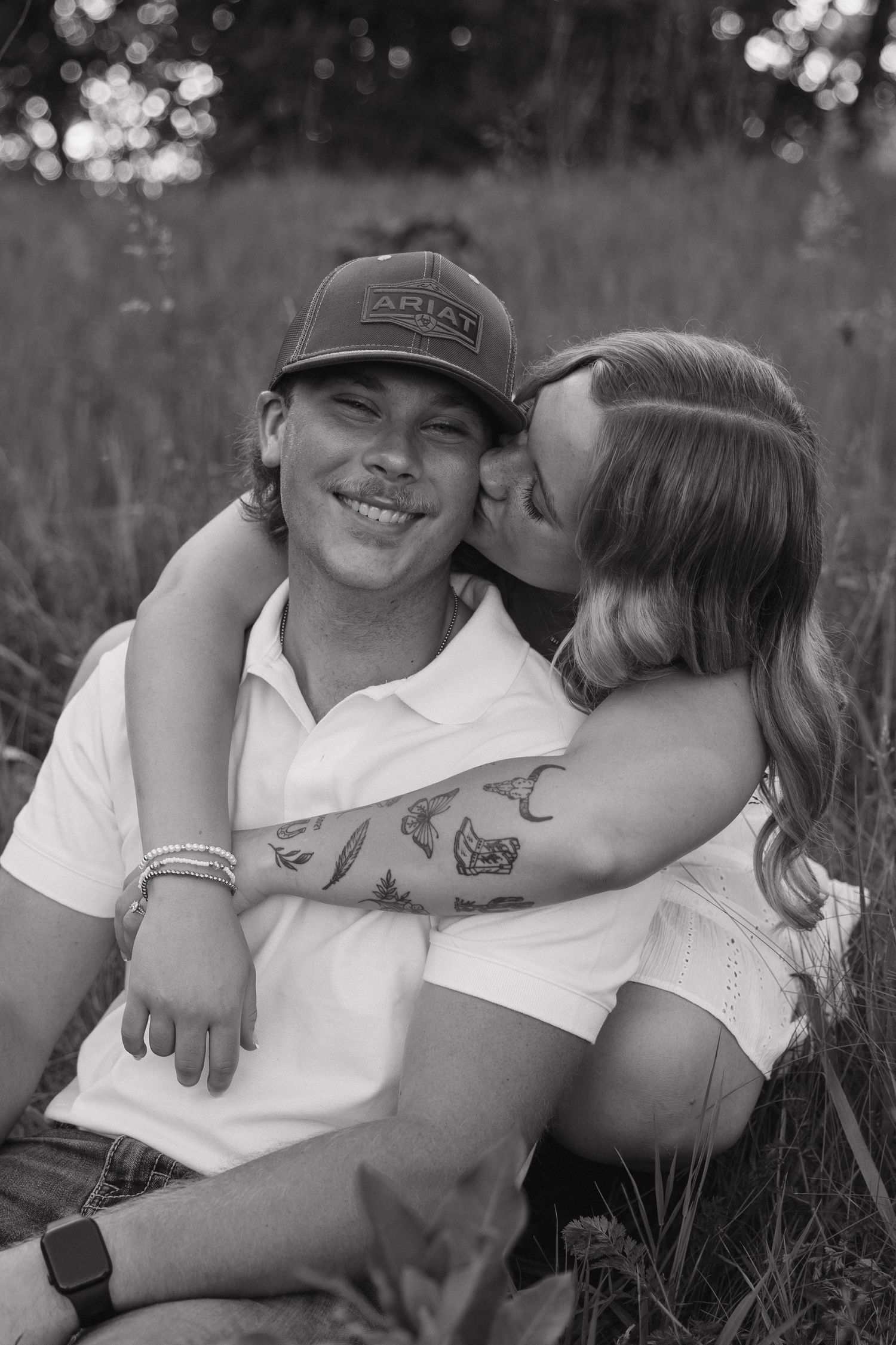Black and white photo of smiling man in Ariat cap being kissed on cheek by woman in grassy field.