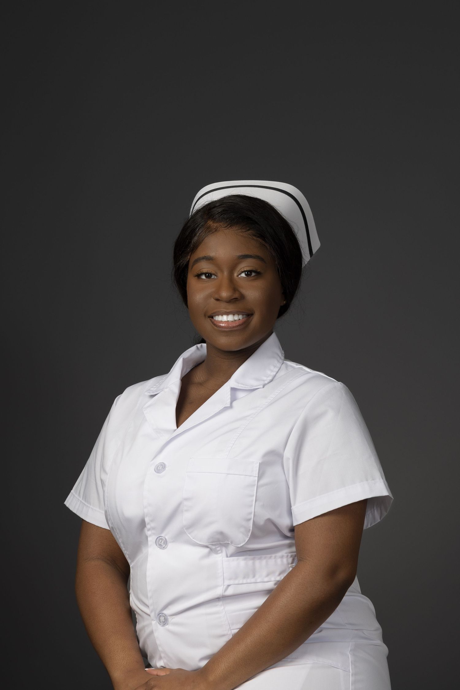 Smiling young Black nurse in white uniform and traditional cap poses confidently against gray background.