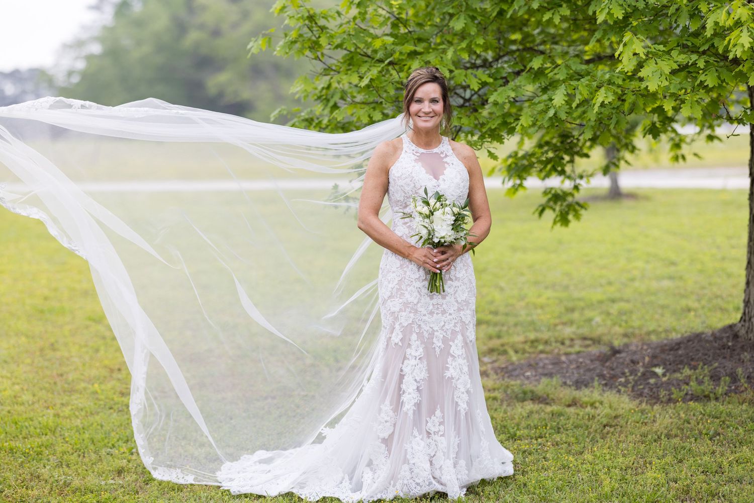 Bride in lace wedding gown with flowing veil holding white bouquet, standing outdoors on green grass near a tree.