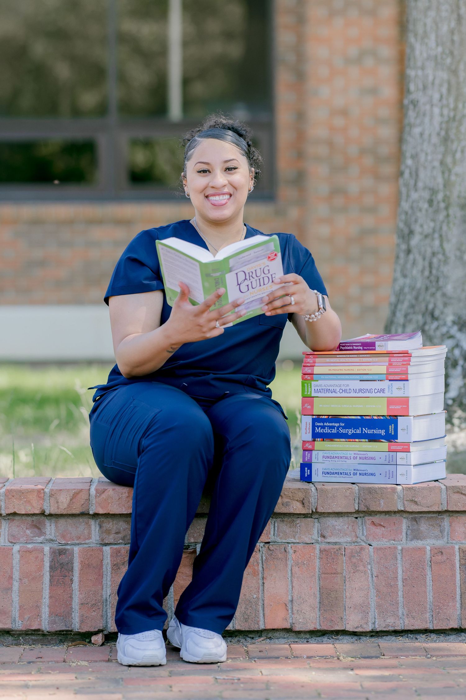Nursing student in blue scrubs smiling while reading a drug guide, seated outdoors with medical textbooks stacked beside her.