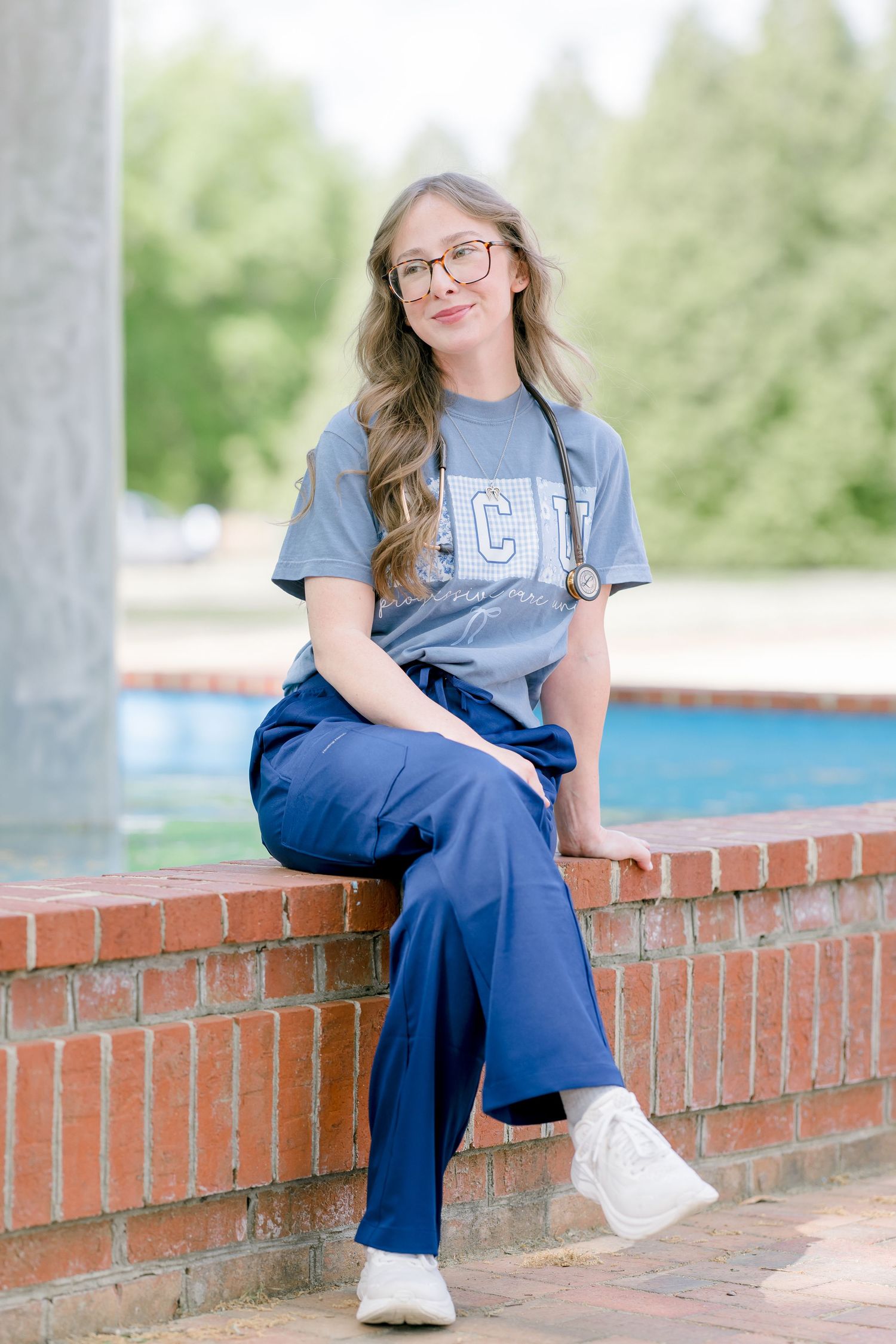 Young woman in blue scrubs and stethoscope sitting on brick wall outdoors, smiling at camera.
