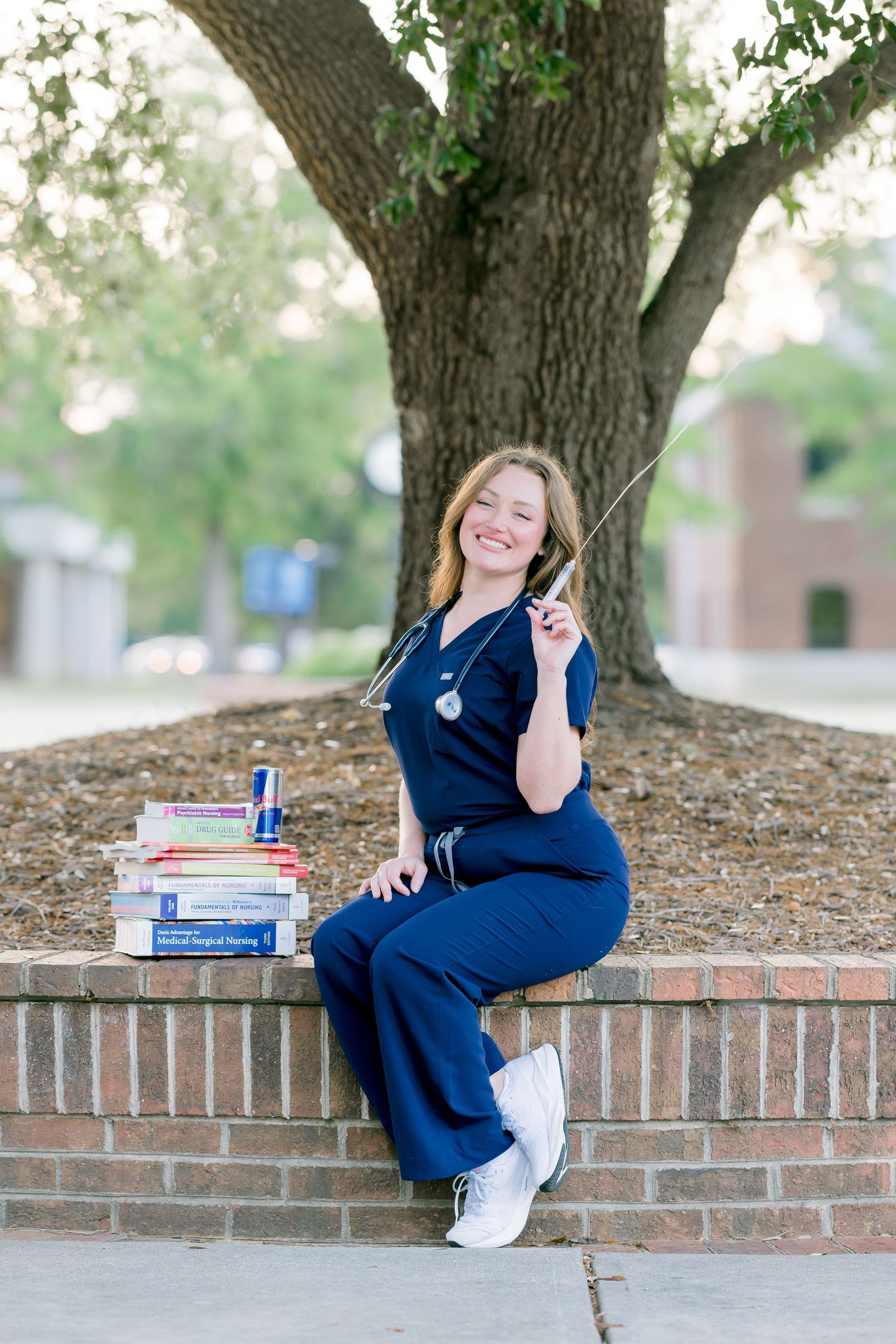Smiling nursing student in navy scrubs sits on brick wall with stethoscope and medical textbooks under large oak tree.