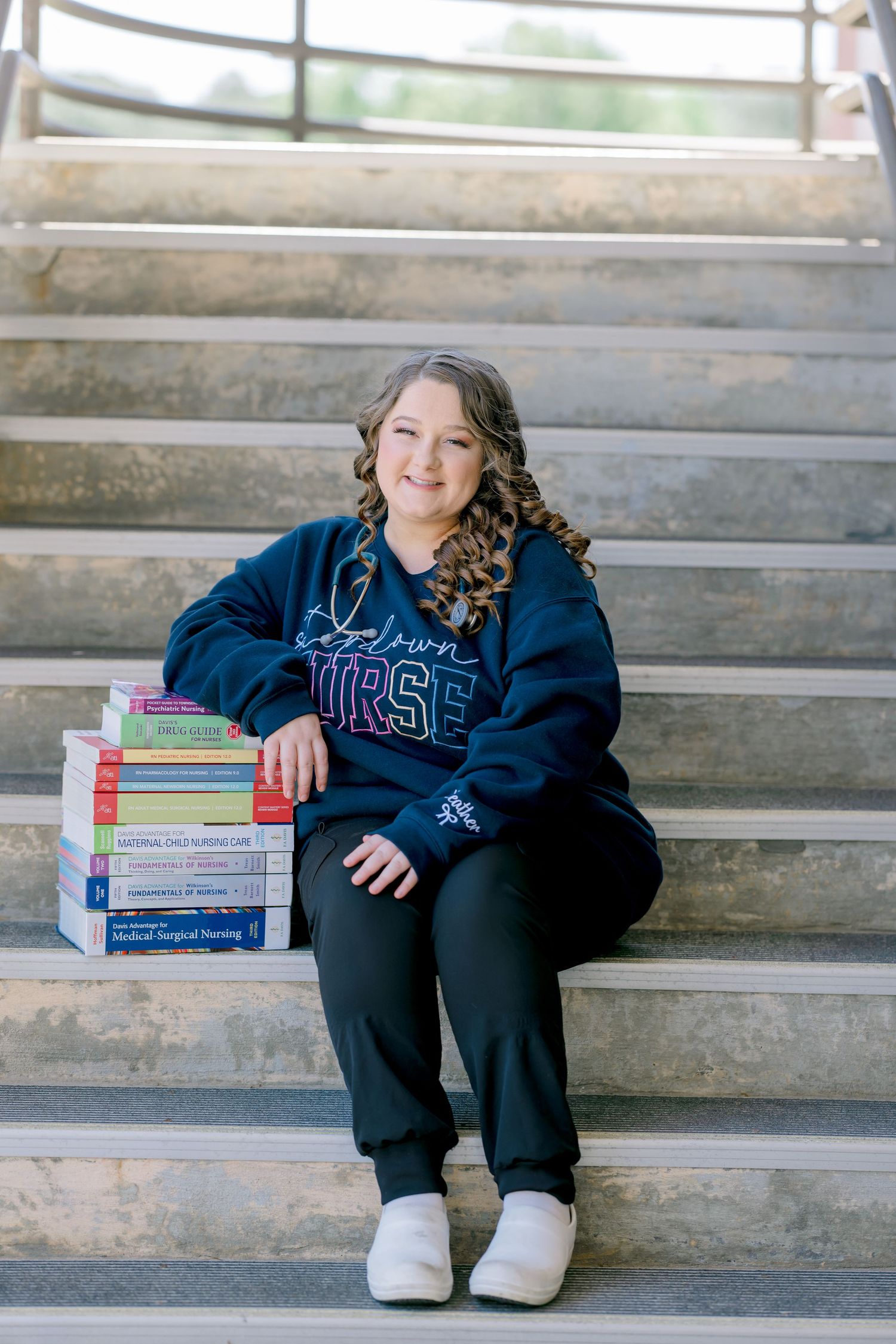 Nursing student sitting on steps smiling with stack of nursing textbooks wearing navy Future Nurse sweatshirt and scrubs.