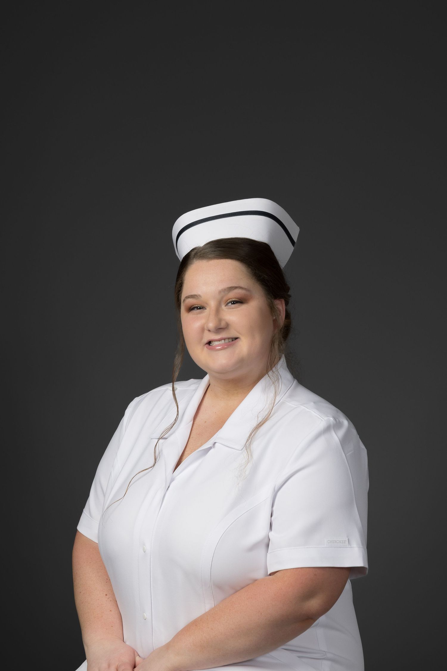 Cheerful nurse in white uniform and cap smiling against gray background.