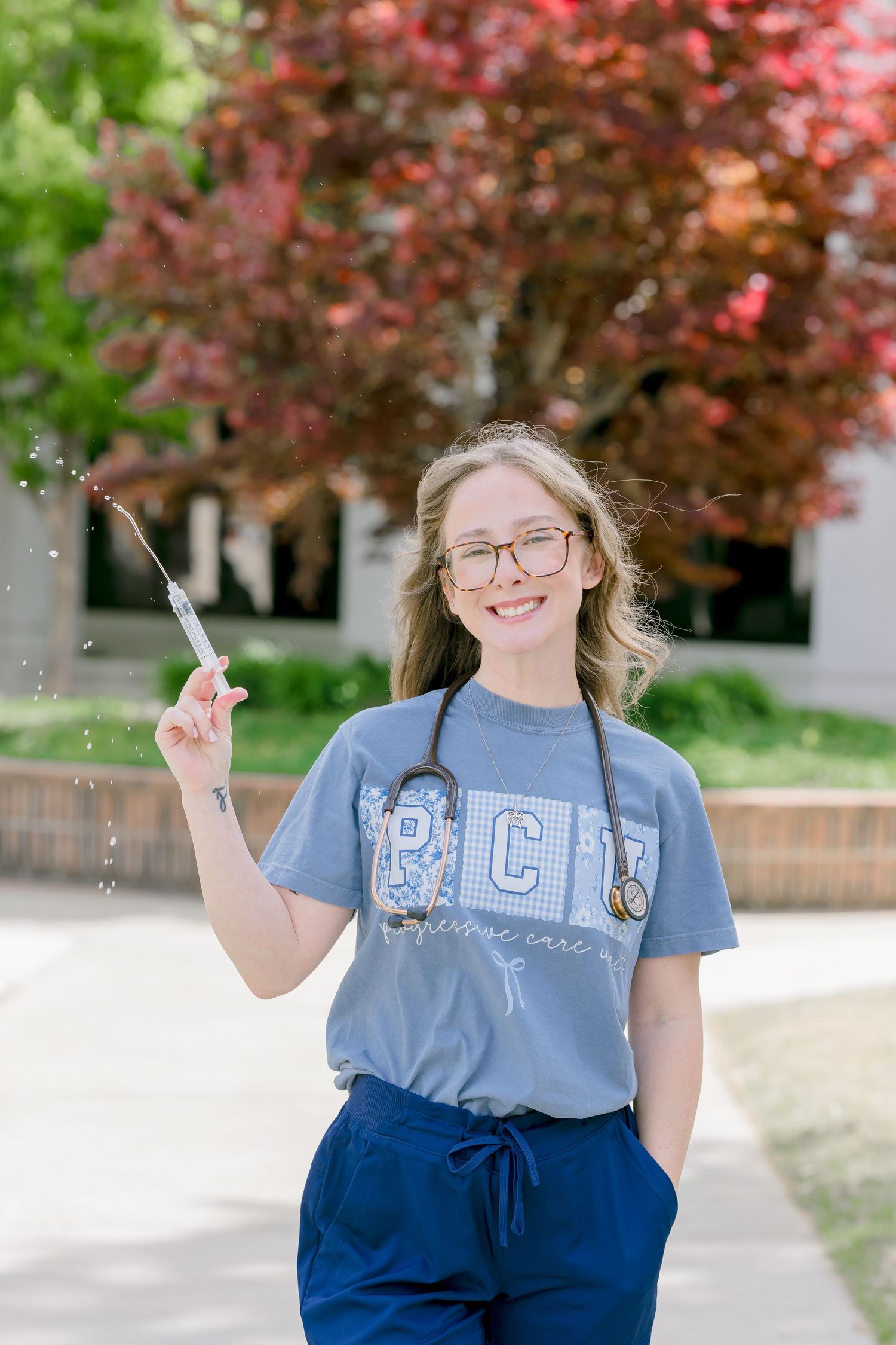 Young nursing student in blue scrubs tossing a syringe in the air while smiling outdoors on campus.