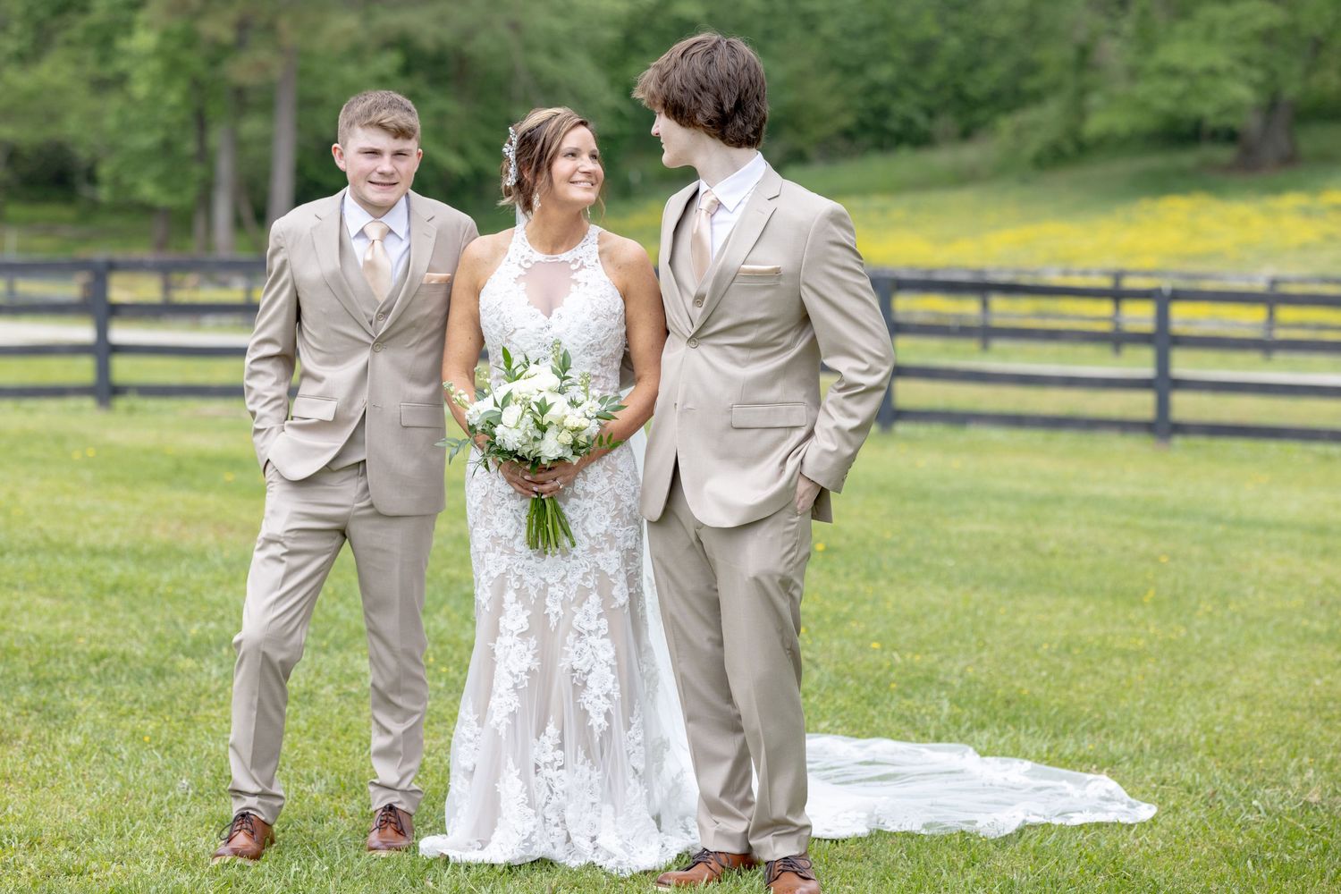 Bride in lace gown holds white bouquet flanked by two groomsmen in tan suits on green lawn near wooden fence.