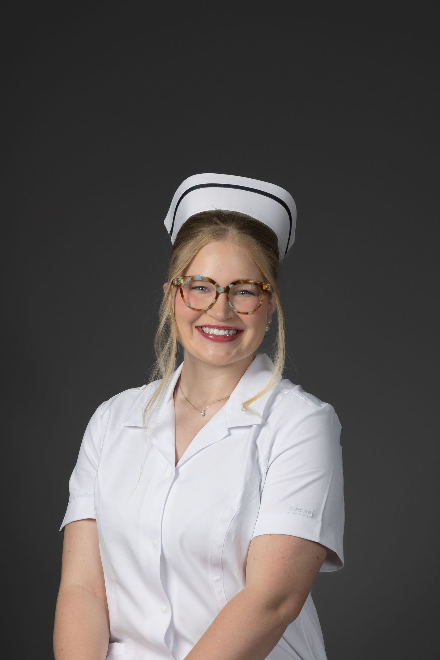 Smiling blonde nurse in white uniform and cap with glasses posing against gray background.