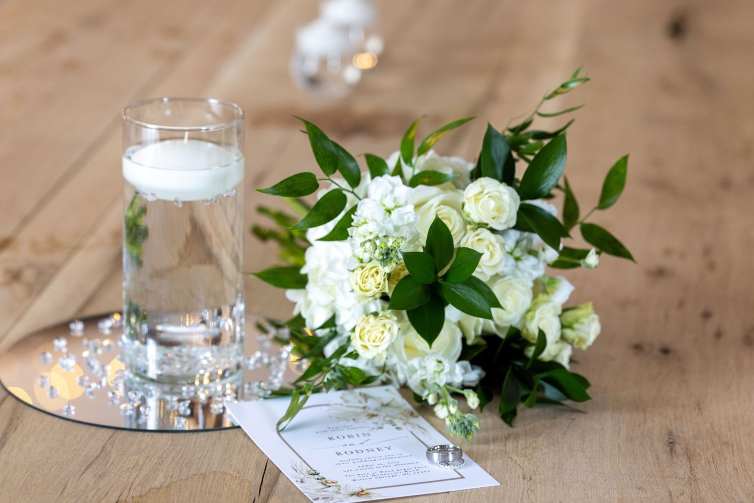 White wedding bouquet with roses and greenery beside a floating candle centerpiece on a wooden table.