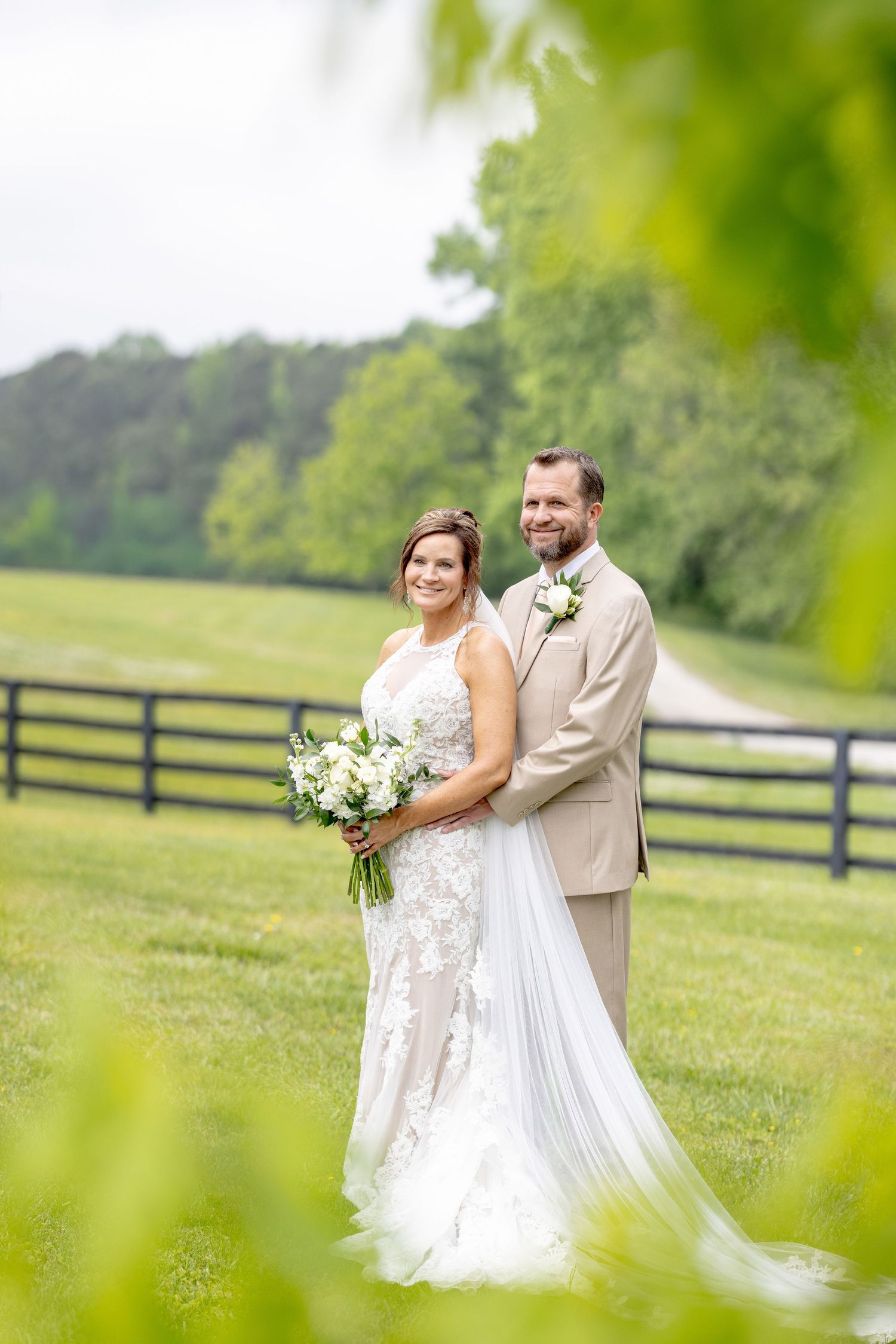 Bride and groom pose together in a lush green field with a wooden fence backdrop on their wedding day.