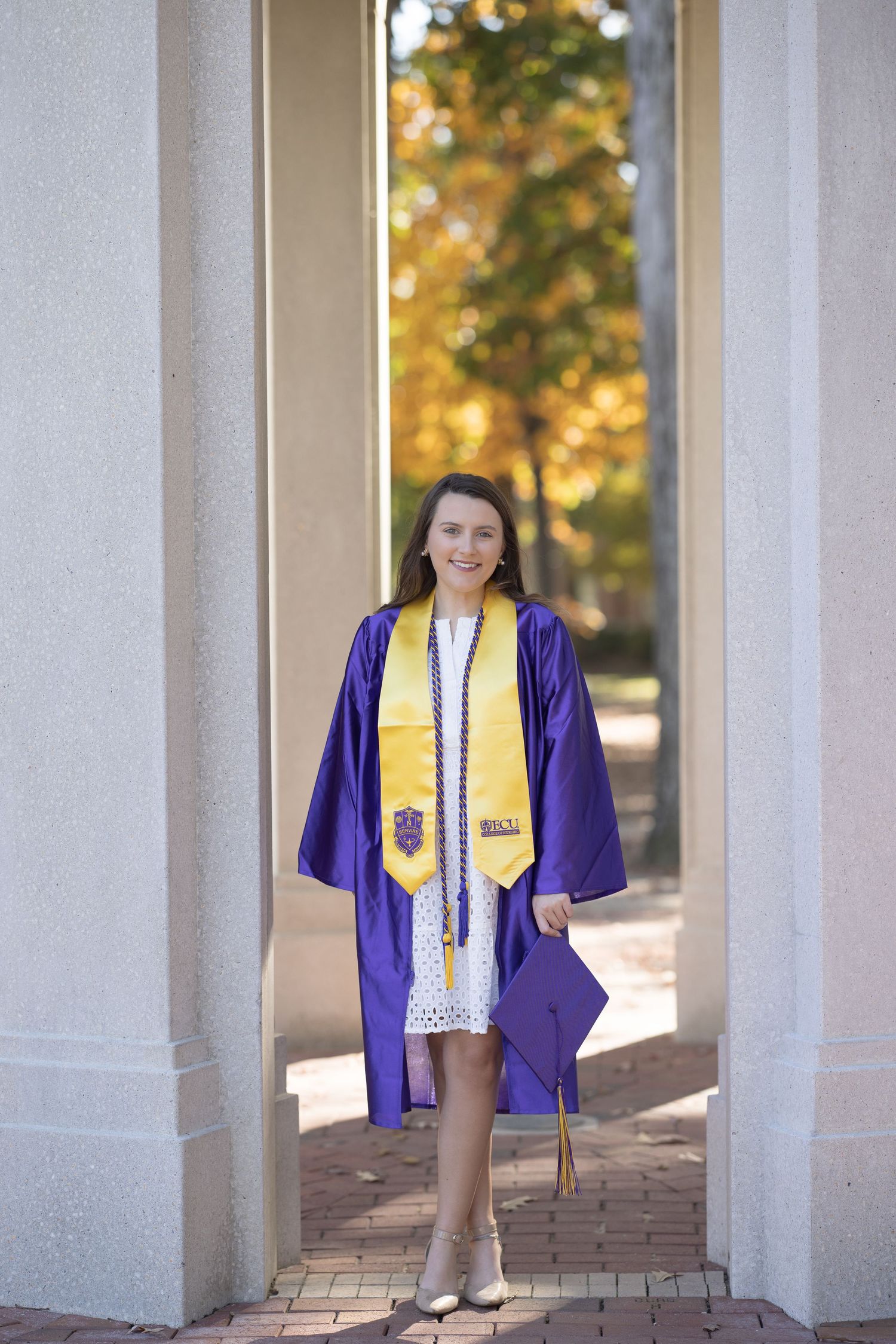 Graduate wearing purple graduation gown and gold stole stands between white columns on sunny autumn day.