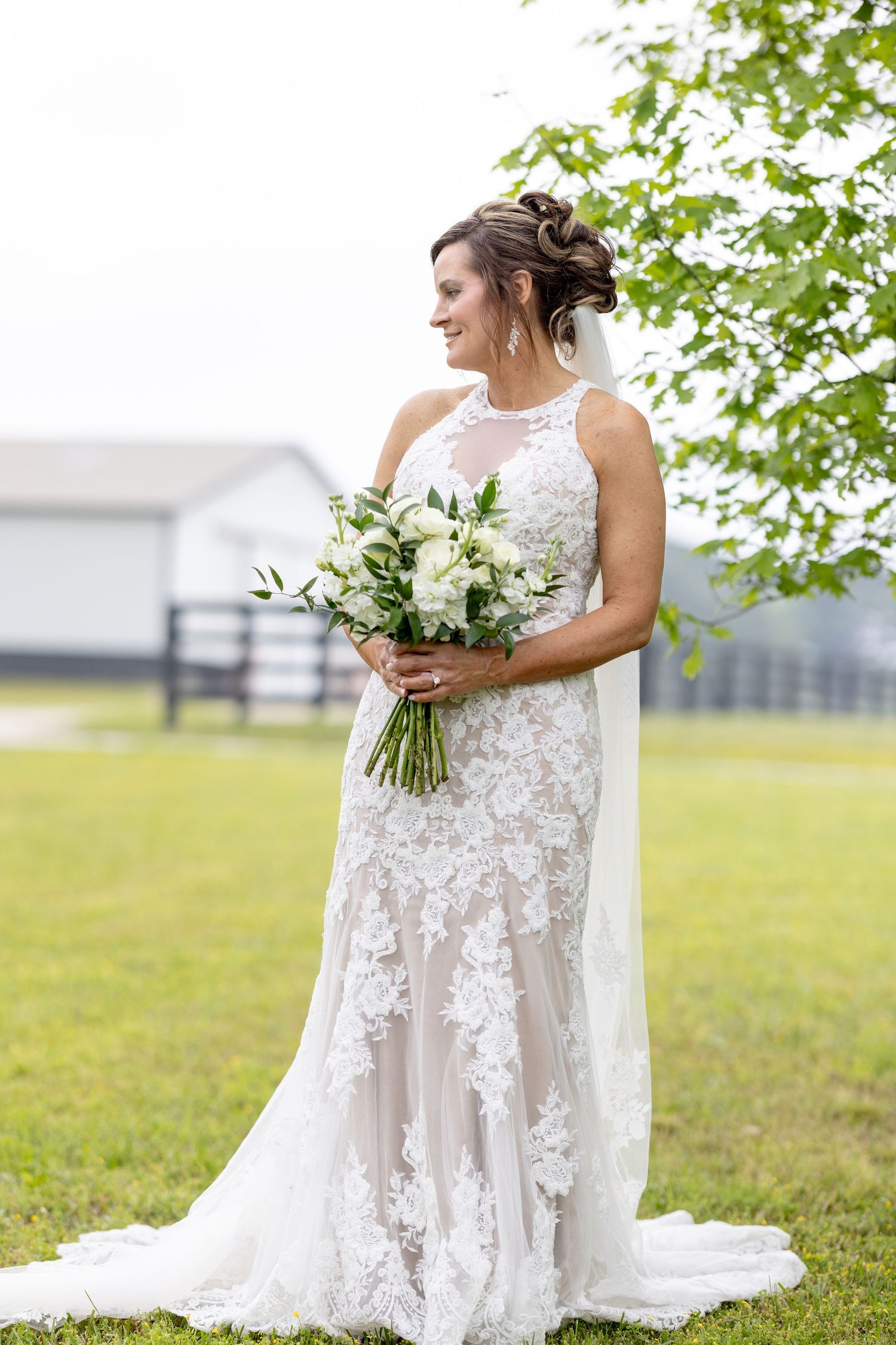 Bride in lace mermaid wedding gown holding white floral bouquet standing outdoors near green tree on wedding day.