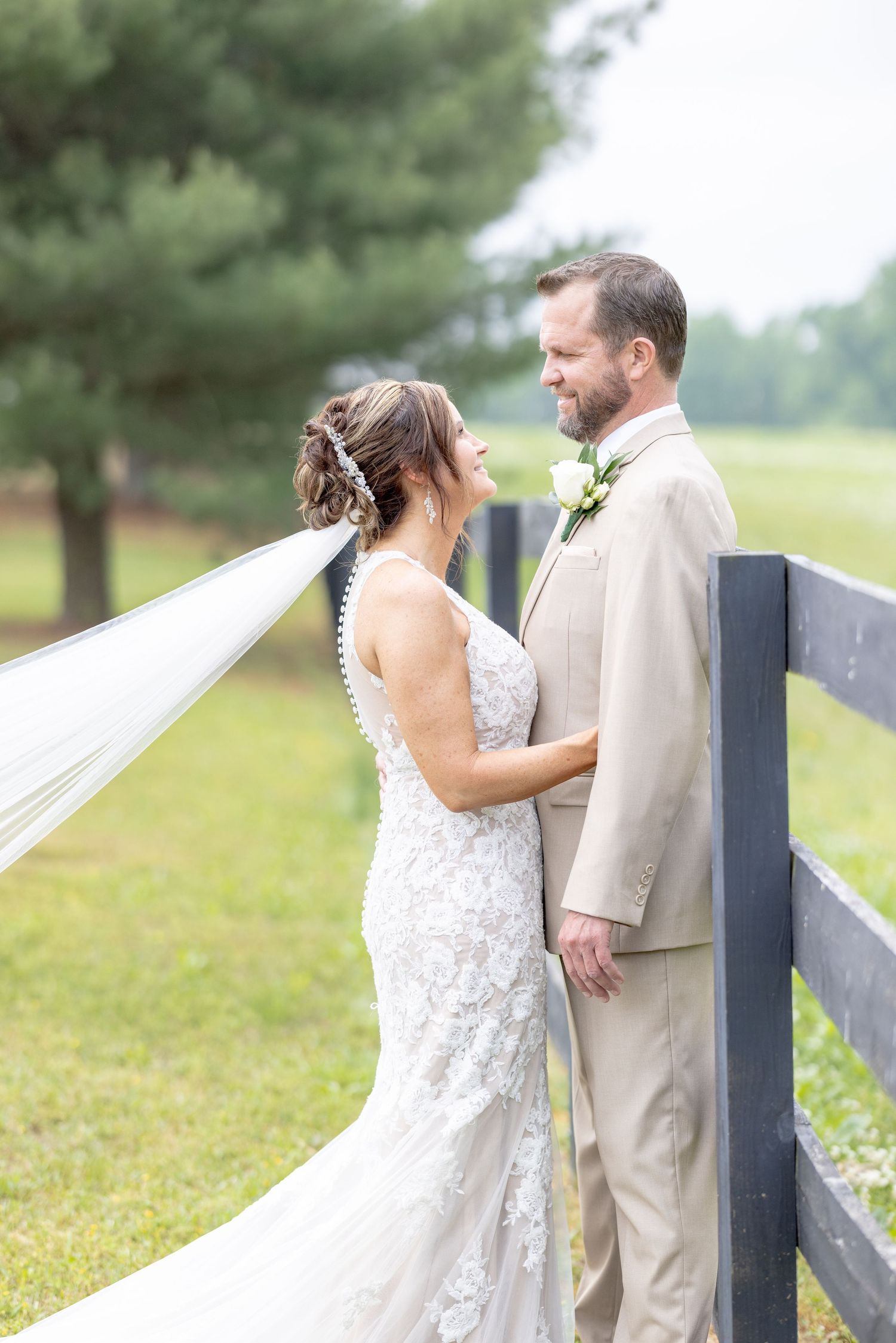 Bride in lace gown and groom in tan suit share a romantic moment by a wooden fence on their wedding day.