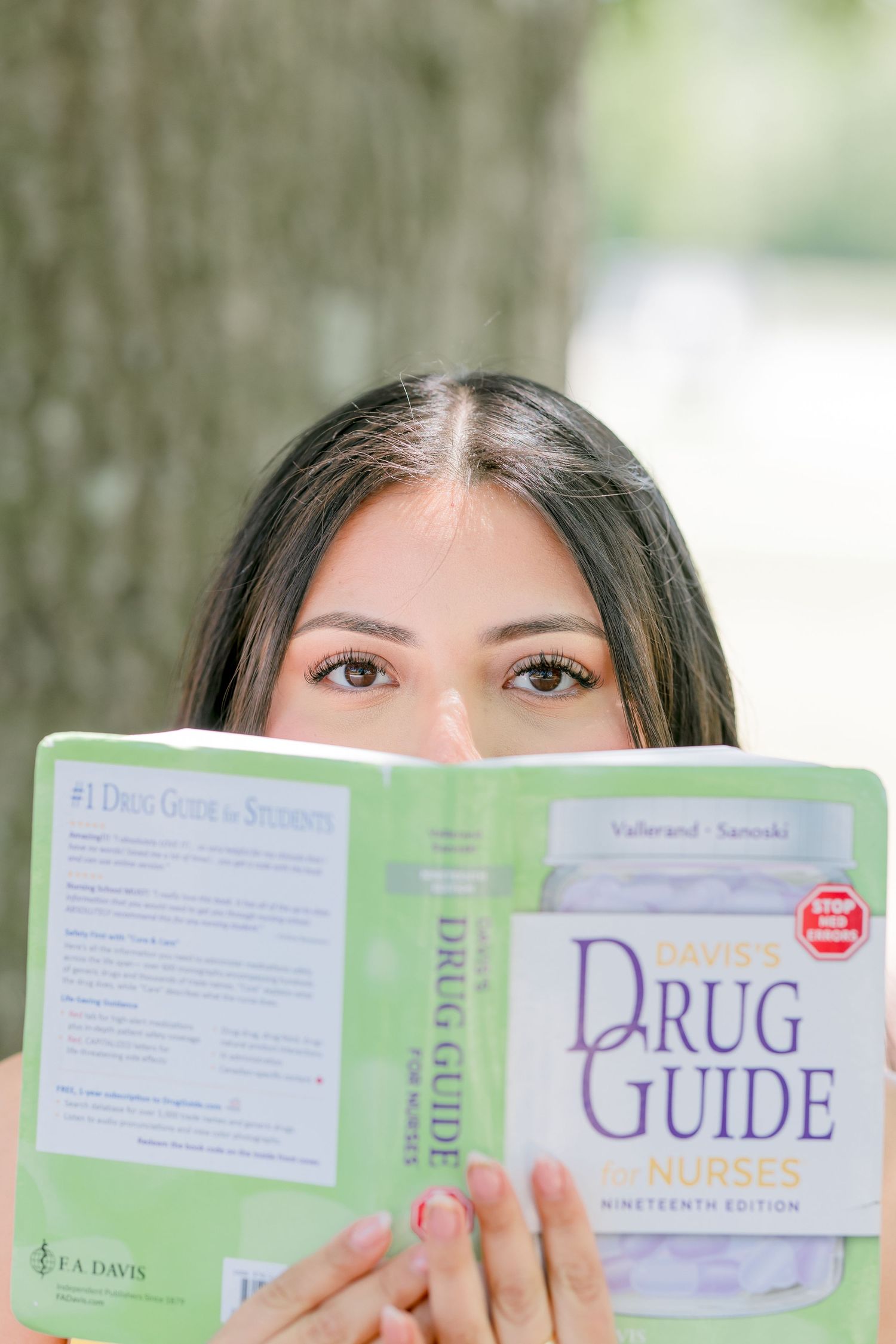 Nursing student holding Davis's Drug Guide for Nurses 19th edition textbook over her face outdoors.