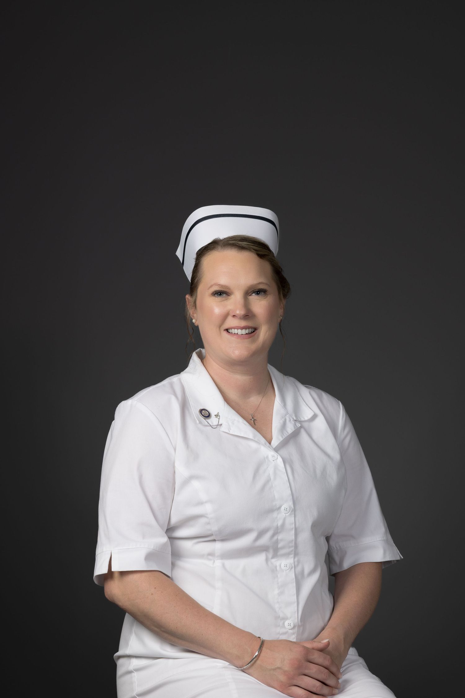 Professional nurse in white uniform and cap smiling confidently against gray background.
