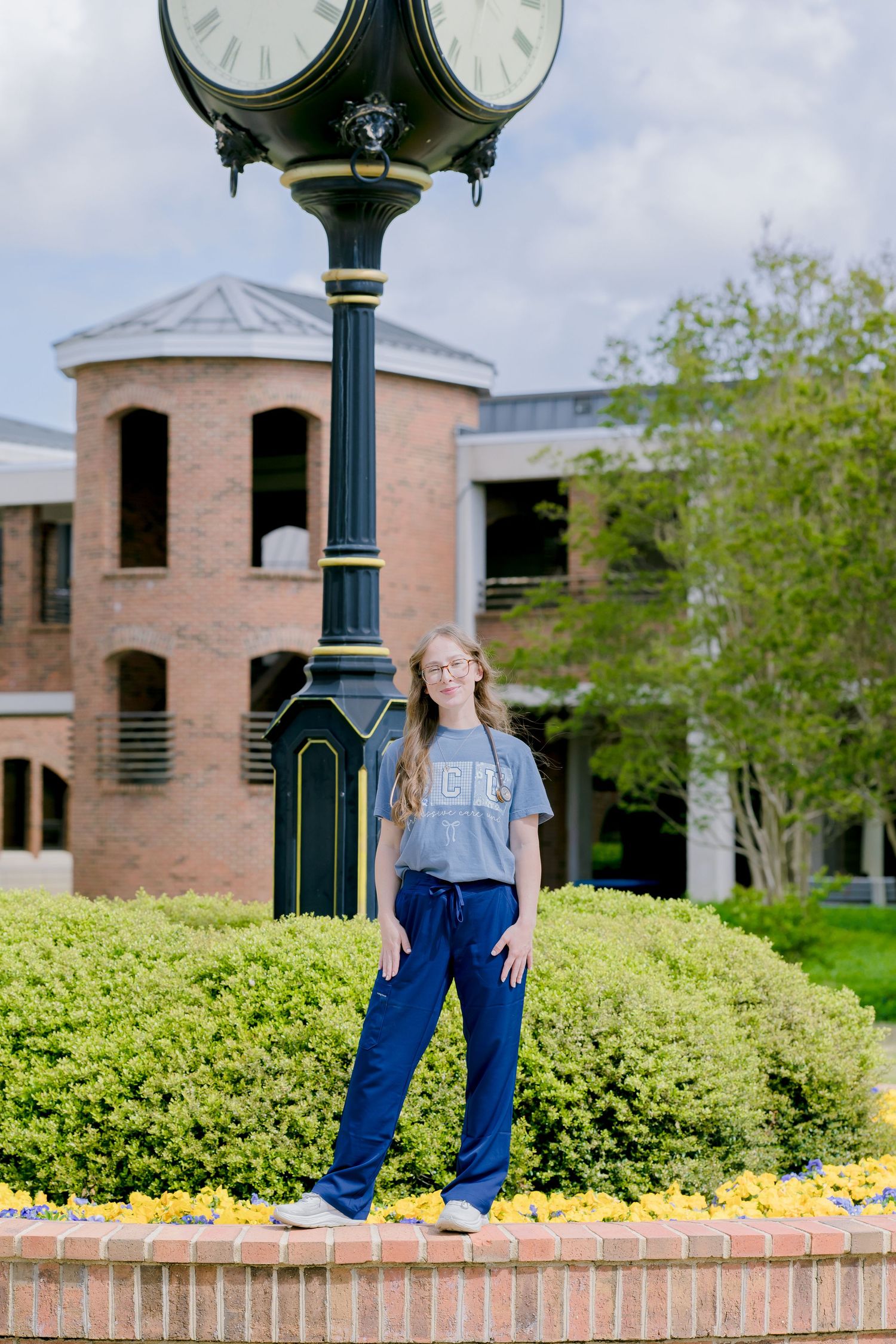 Young woman in gray tee and navy pants poses by ornate street clock near brick building with lush green shrubs.