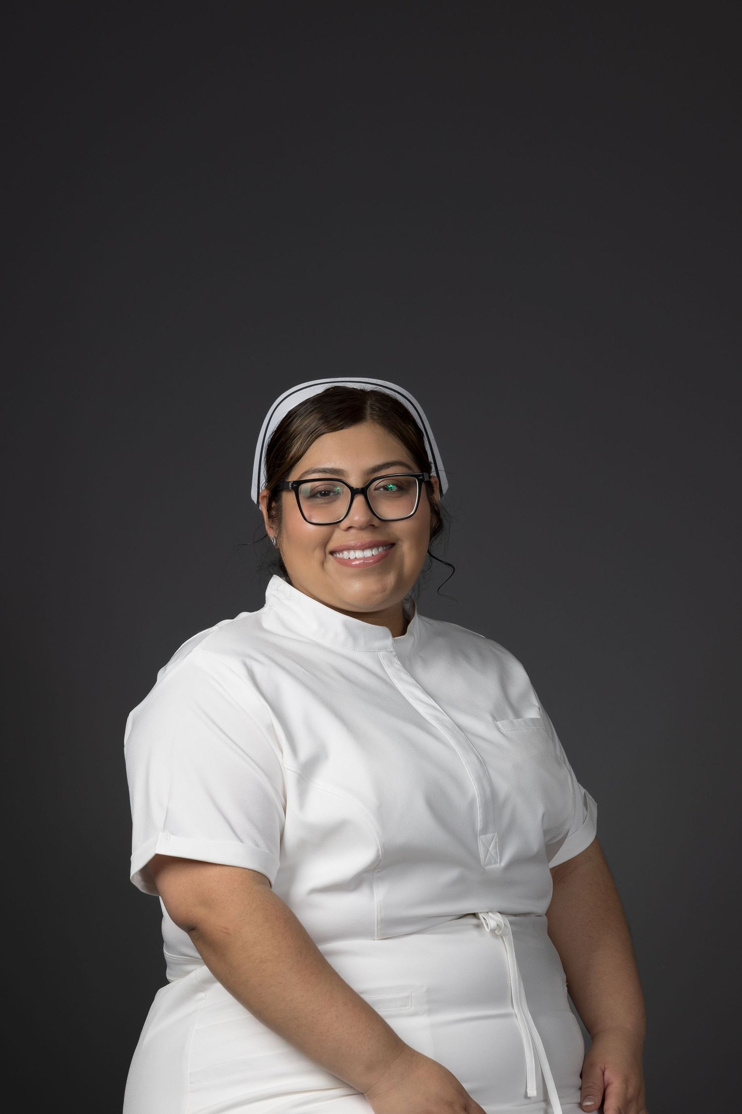 Female nurse wearing glasses and white uniform with cap smiles against gray background.