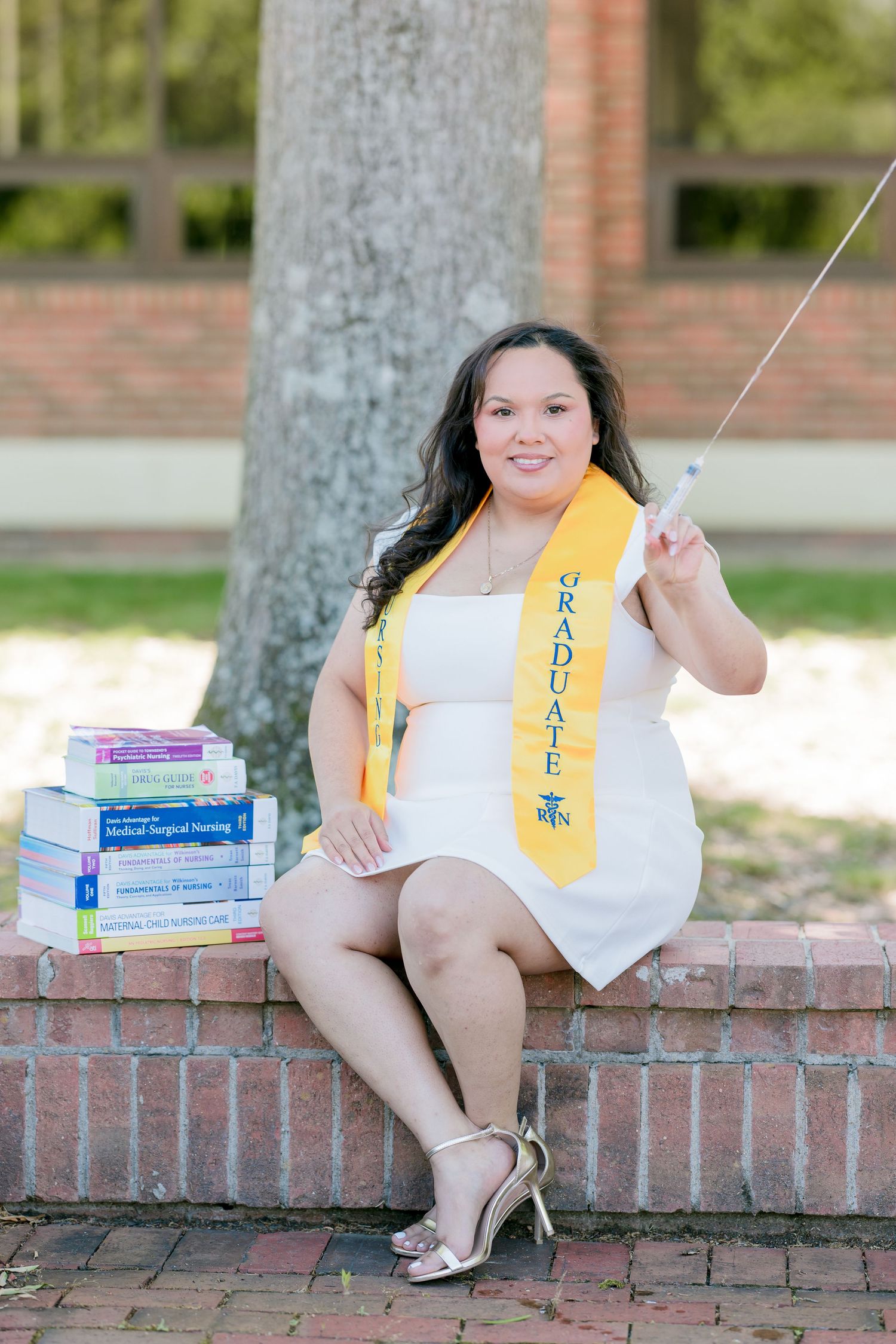 Nursing graduate in white dress and gold sash sits on brick wall with medical textbooks, holding a syringe.