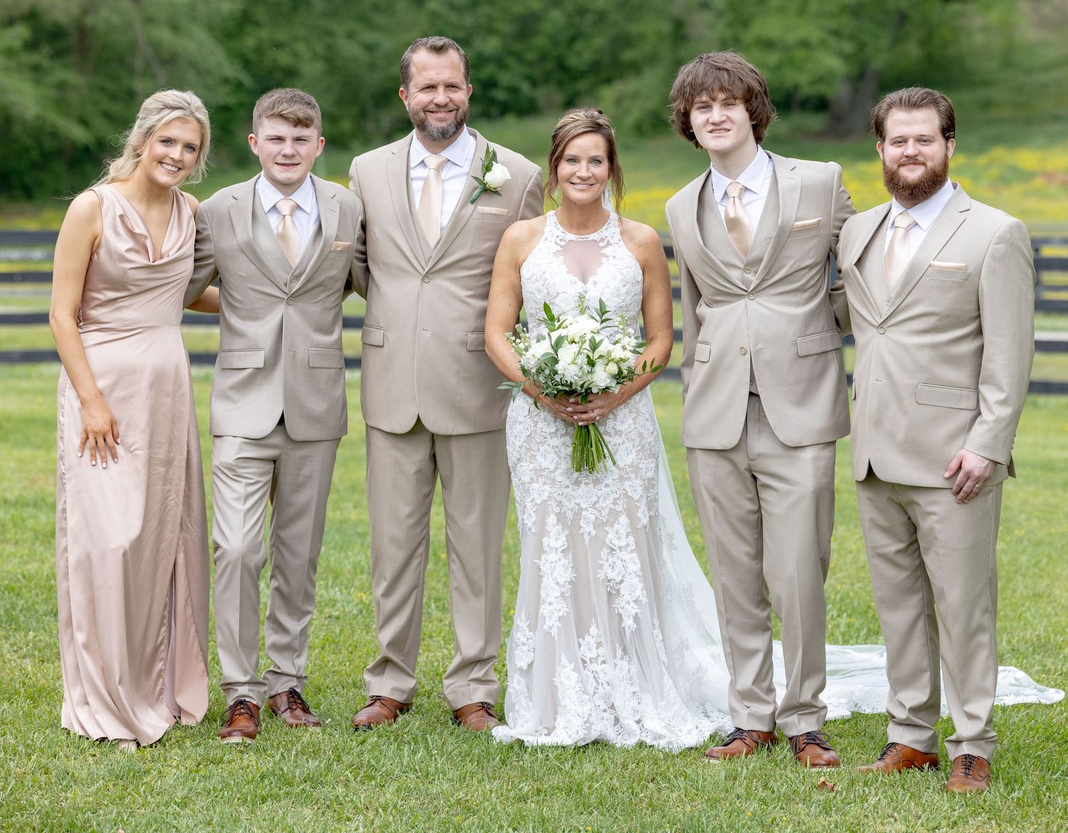 Wedding party posing outdoors, bride in lace gown with bouquet, groomsmen and bridesmaid in tan and blush attire.