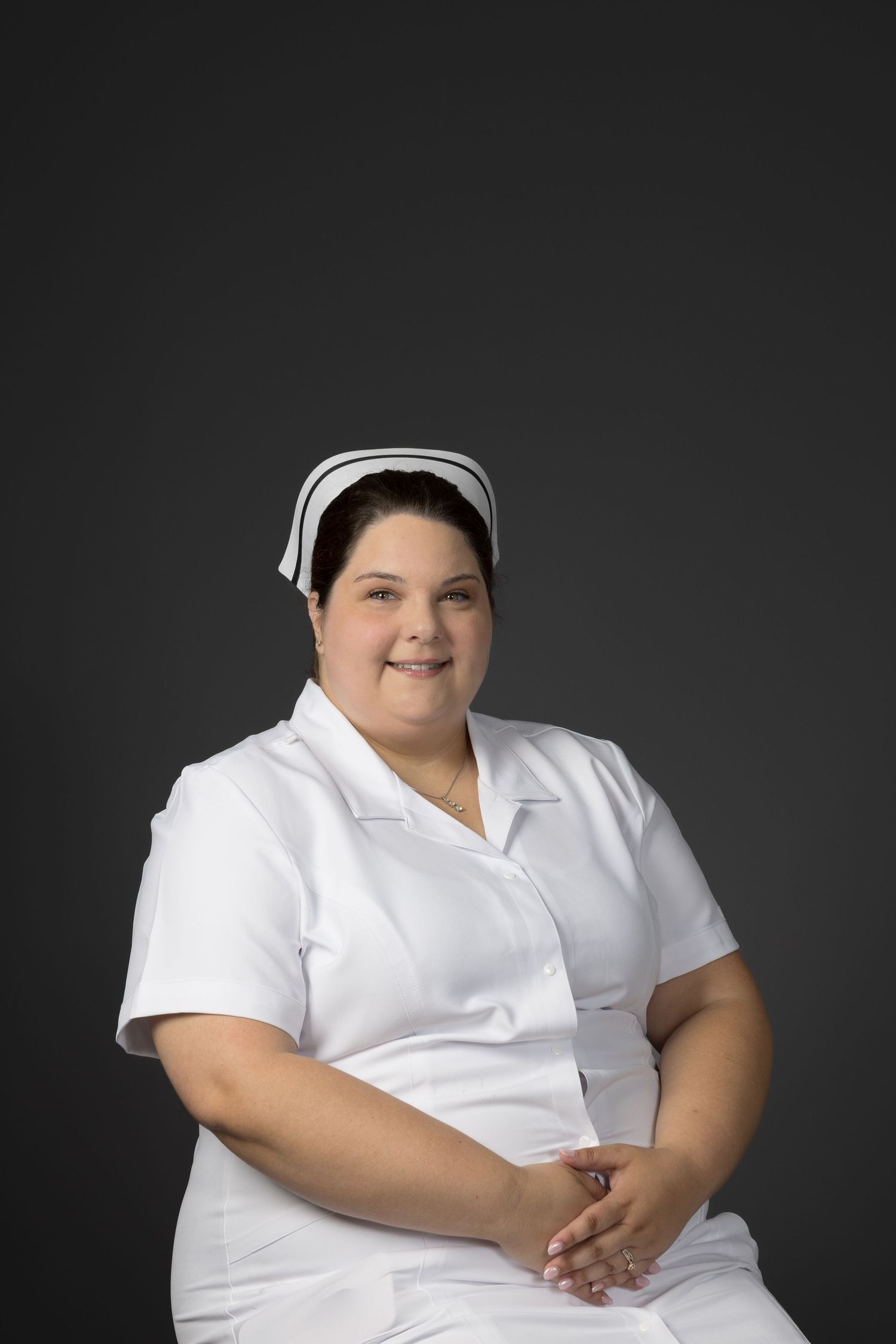 Plus-size female nurse in white uniform and cap smiling against gray background.