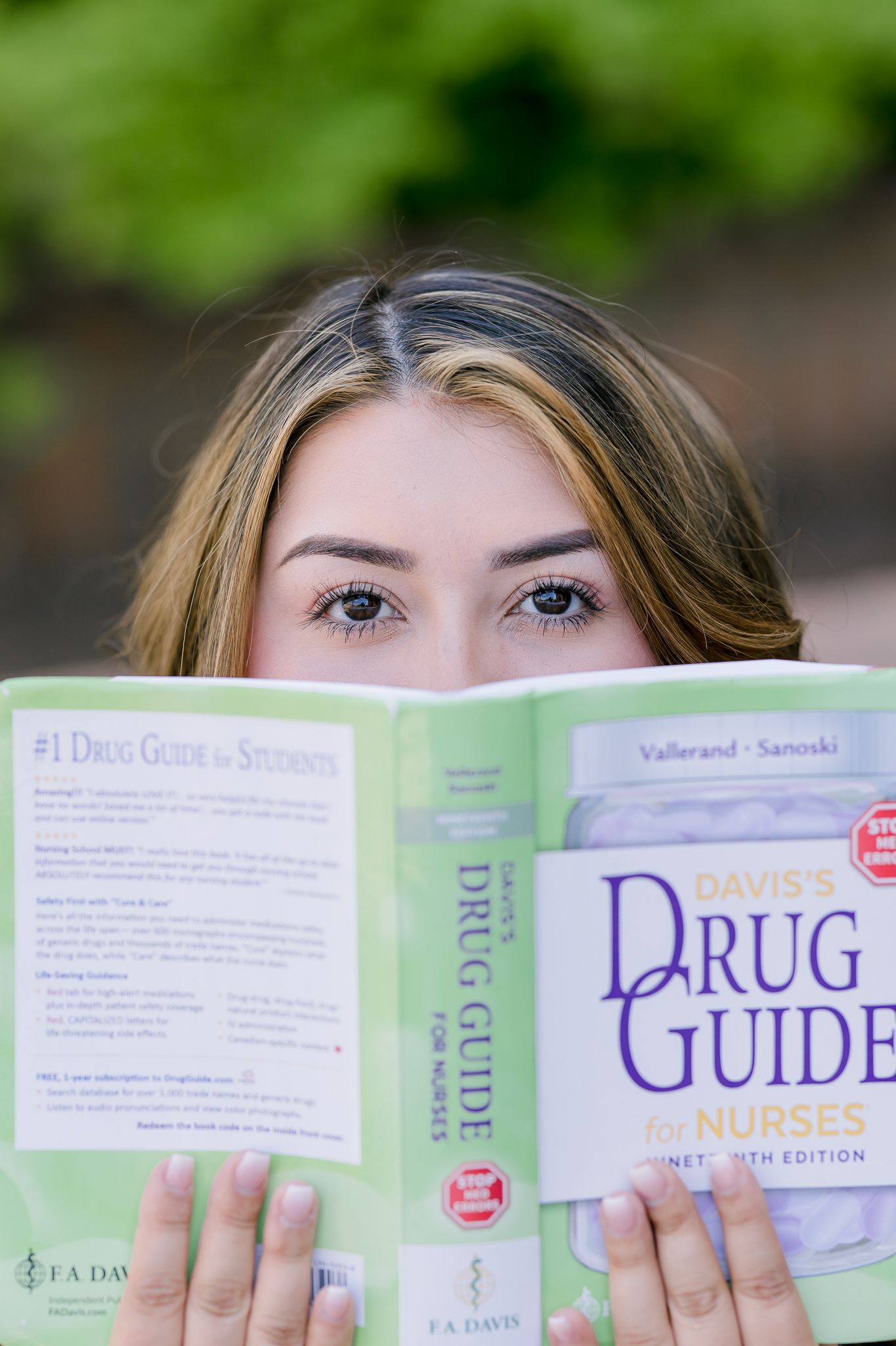 A nursing student holds Davis's Drug Guide for Nurses textbook up to her face outdoors.