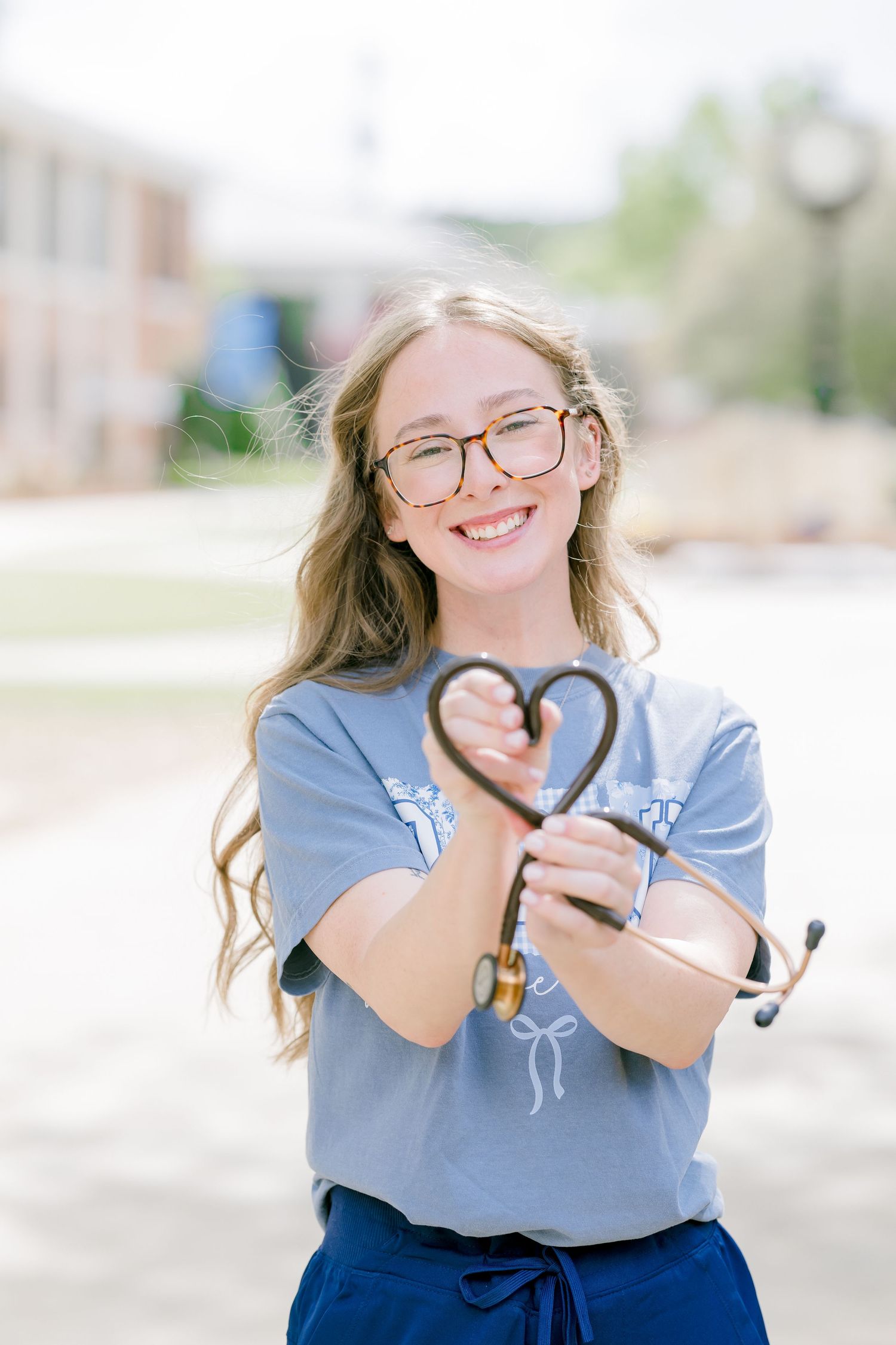 Smiling nursing student holding stethoscope in heart shape outdoors, wearing glasses and blue scrub top.