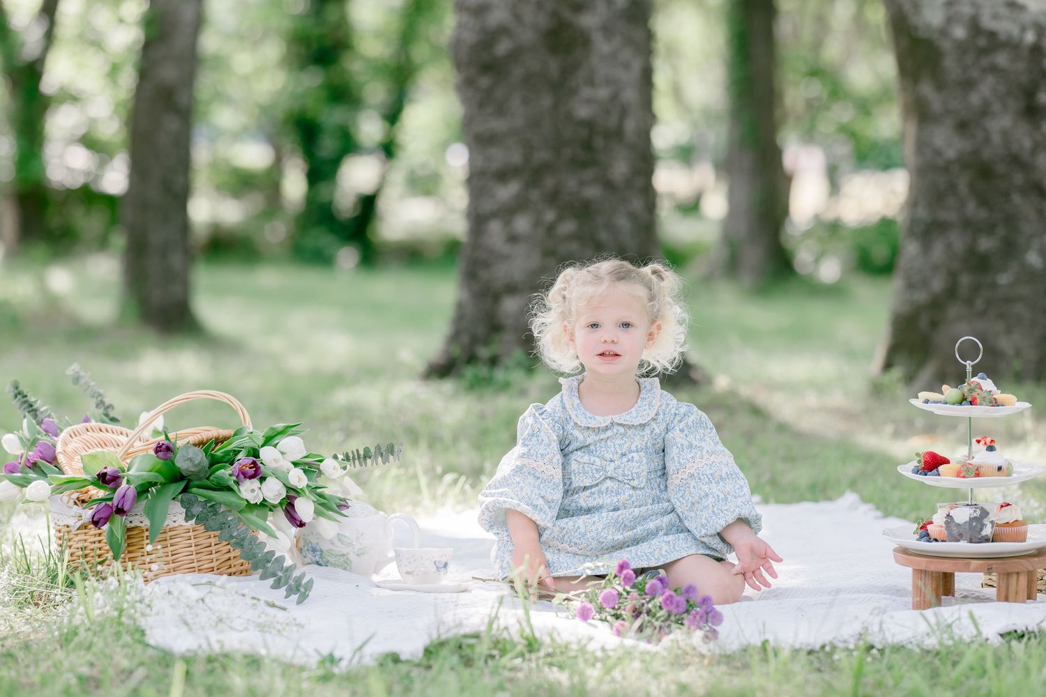 Toddler girl in gray dress sits on white blanket at outdoor picnic with flowers and treats in green park.