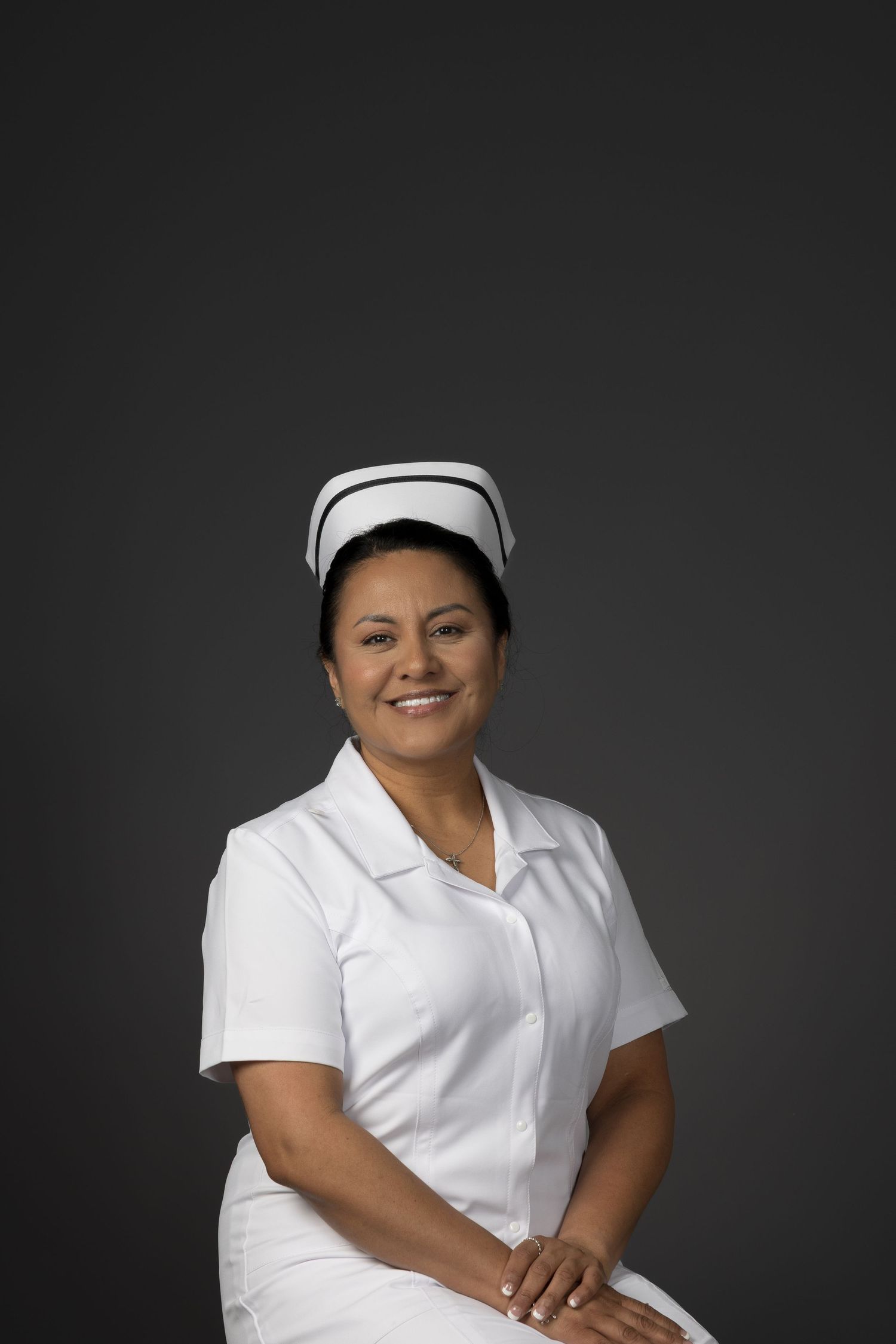 Smiling woman in white nurse uniform and cap seated against dark gray studio background.